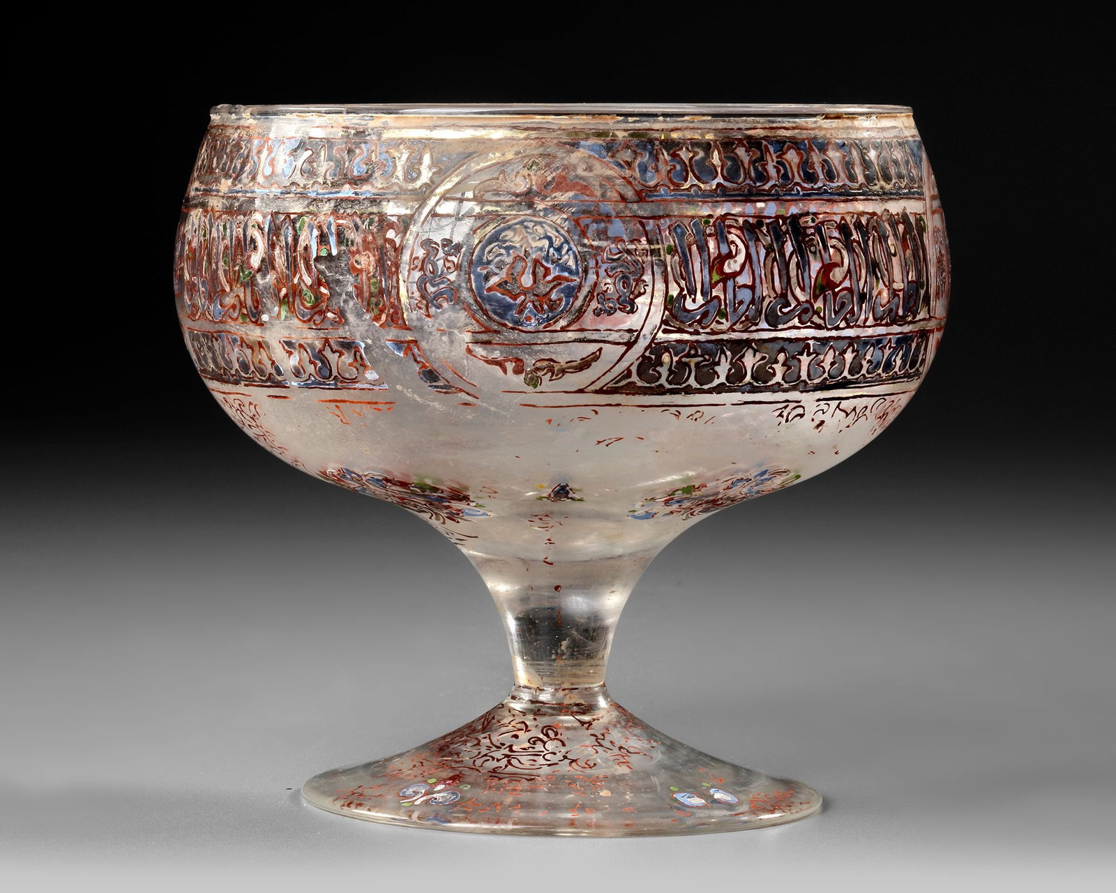 AN ISLAMIC GILDED AND ENAMELED FOOTED BOWL, 19TH-20TH CENTURY: An Islamic glass footed bowl: a rounded bowl with conical sides, standing on a pronounced, tall foot. It is enamelled in red, green, blue, and traces of gilding. The bowl features a wide band containi