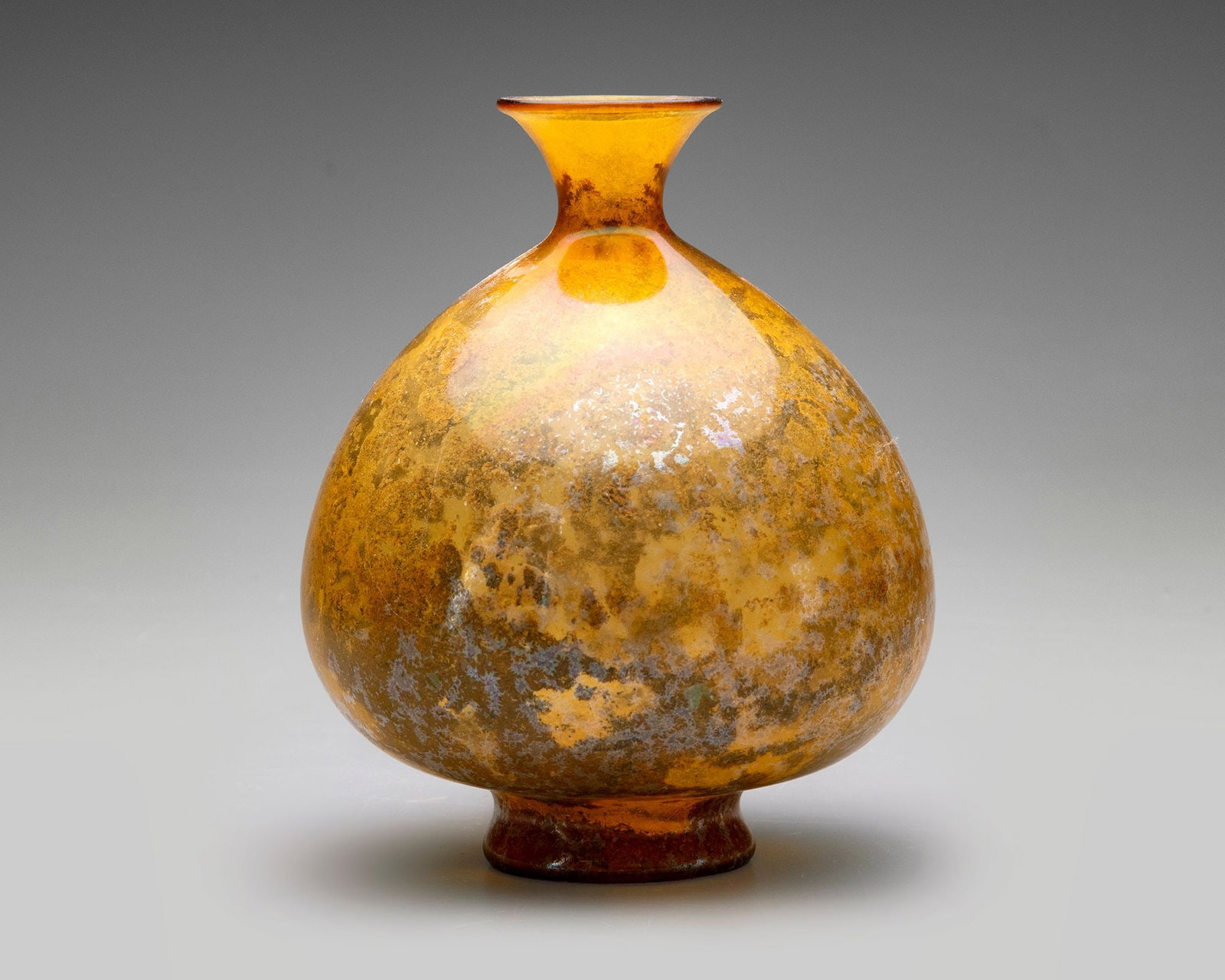 AN AMBER GLASS VESSEL, PERSIA, 10THâ€“11TH CENTURY: Amber-colored glass vessel with a rounded body and short, flaring neck. The surface shows heavy weathering and iridescence. Height 14 cm.