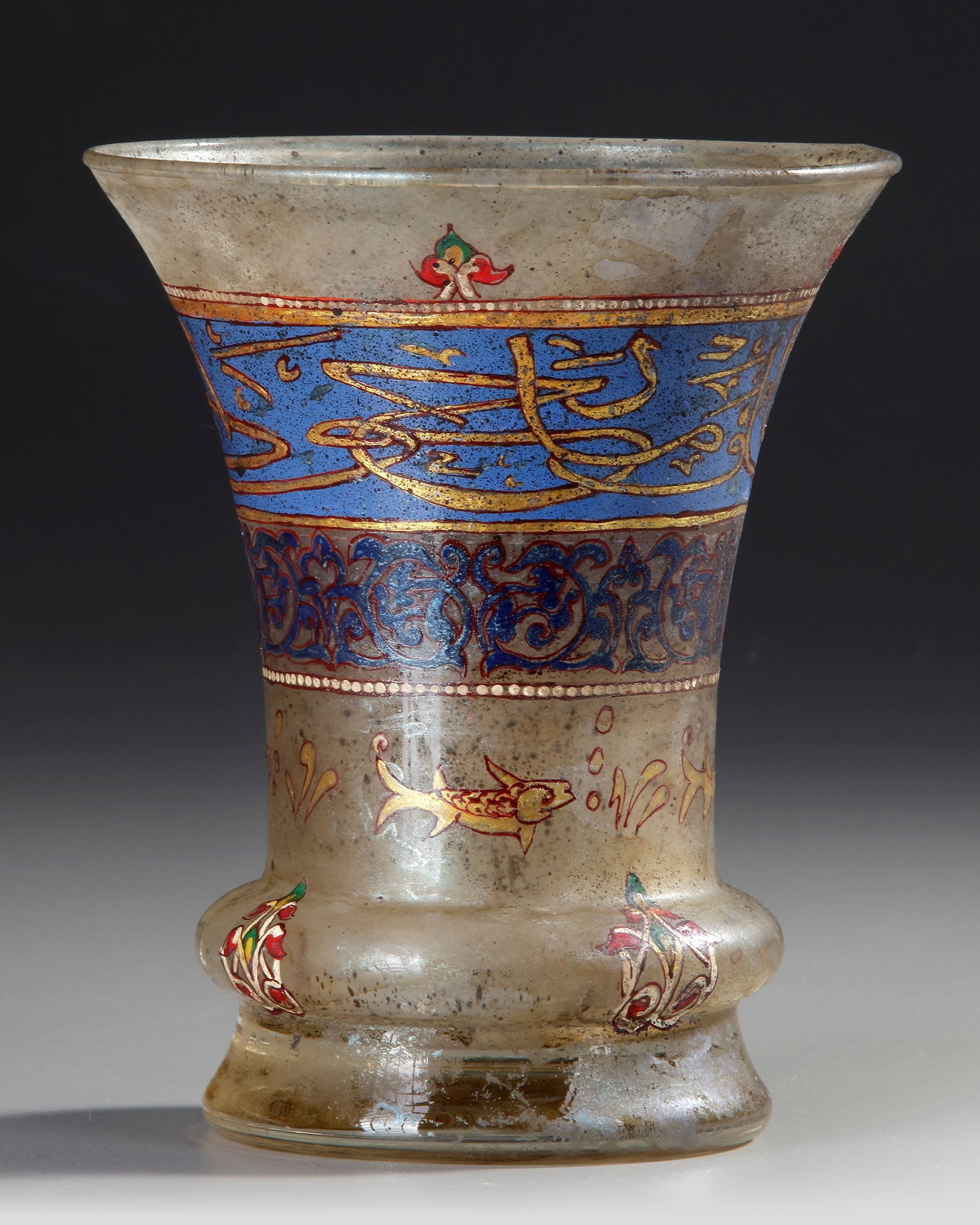 AN ISLAMIC ENAMELLED GLASS BEAKER, 19TH CENTURY: Of cylindrical form with flaring mouth, kick to base, decorated in polychrome enamel and gilt with a band of inscriptions in thuluth script on a blue ground. 20 by 15 cm.