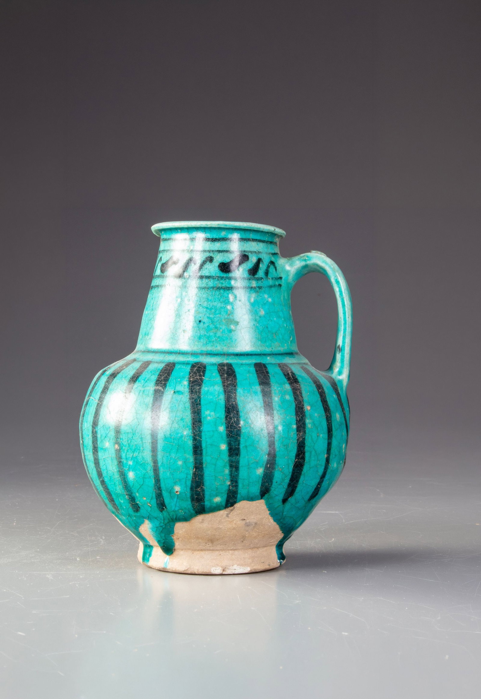 AN UNDERGLAZE PAINTED POTTERY EWER, 12THâ€“13TH CENTURY: A finely crafted Syrian pottery ewer decorated with vibrant turquoise glaze and bold black underglaze stripes. The rounded body, elegant handle, and flowing glaze. Height 16 cm.