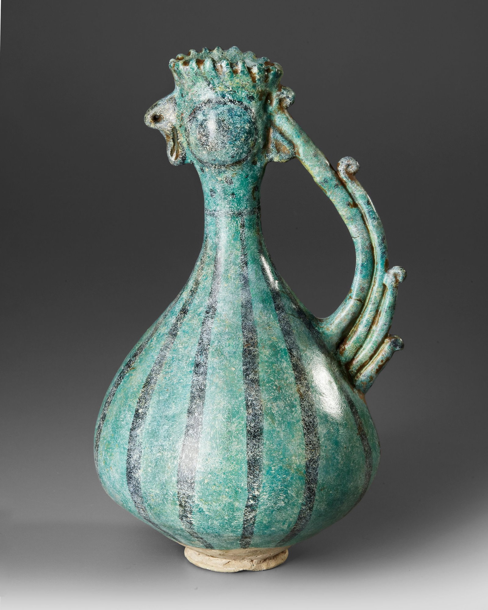 A KASHAN TURQUOISE GLAZED COCKEREL-HEAD POTTERY EWER, PERSIA, 13TH CENTURY: A pottery ewer with a spherical body and tubular neck, the mouth formed as a cockerelâ€™s head with a flaring comb. The handle takes the form of a rising, tapering, curving tail. The vessel is