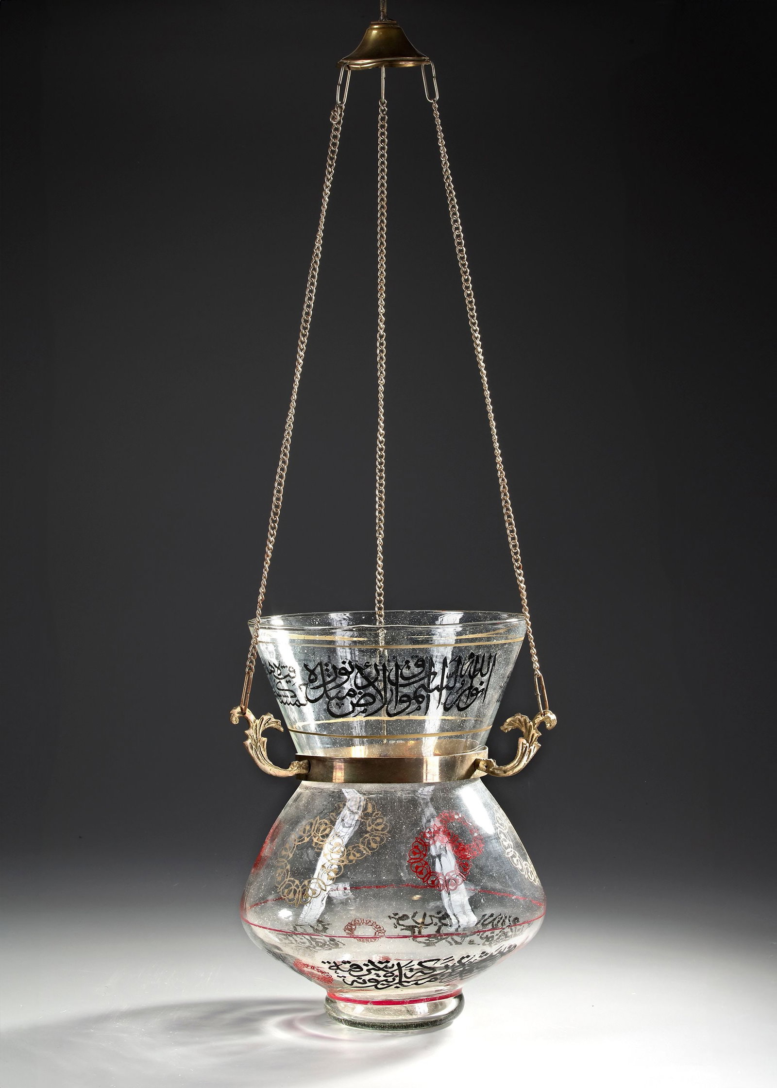 A MAMLUK-STYLE GLASS MOSQUE LAMP, 20TH CENTURY: A glass mosque lamp with a large round bulbous body rising to a narrower waist, standing on a foot so it can be placed on a surface, but suspended by chains, decorated with floral roundels and Inscrip