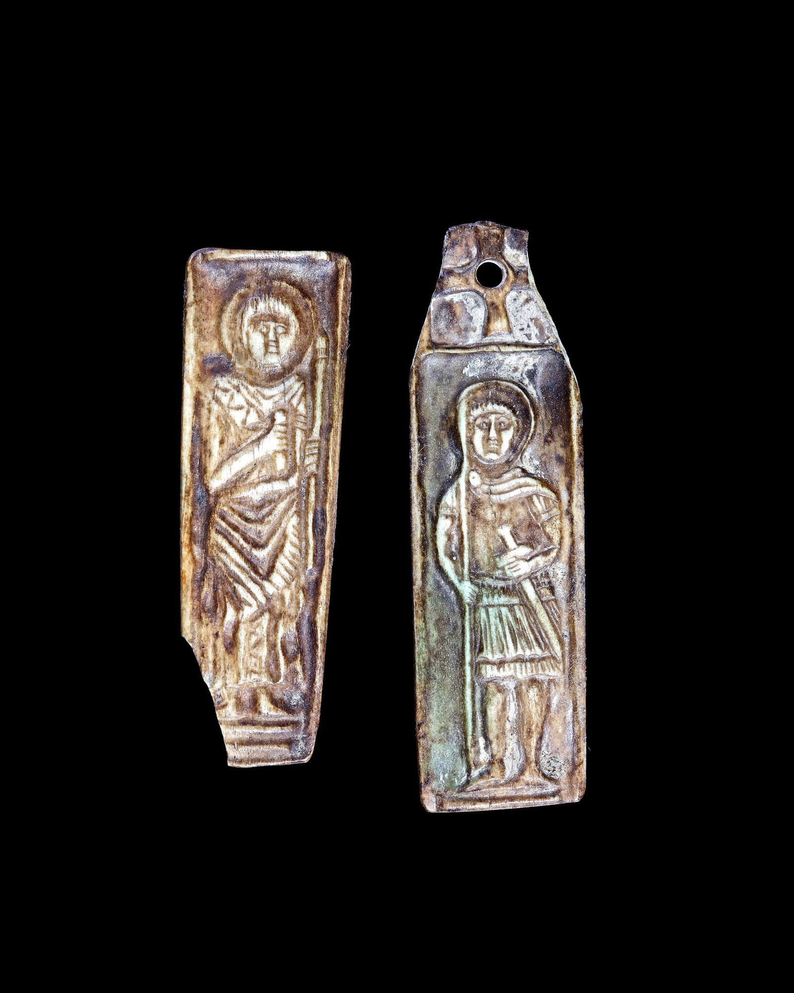 TWO BYZANTINE BONE PLAQUES DEPICTING SACRED FIGURES, 10THâ€“11TH CENTURY: Two Byzantine carved bone plaques, each decorated with a standing haloed sacred figure. One figure holds a staff, while the other is depicted in military attire holding a spear and sword. The plaques 