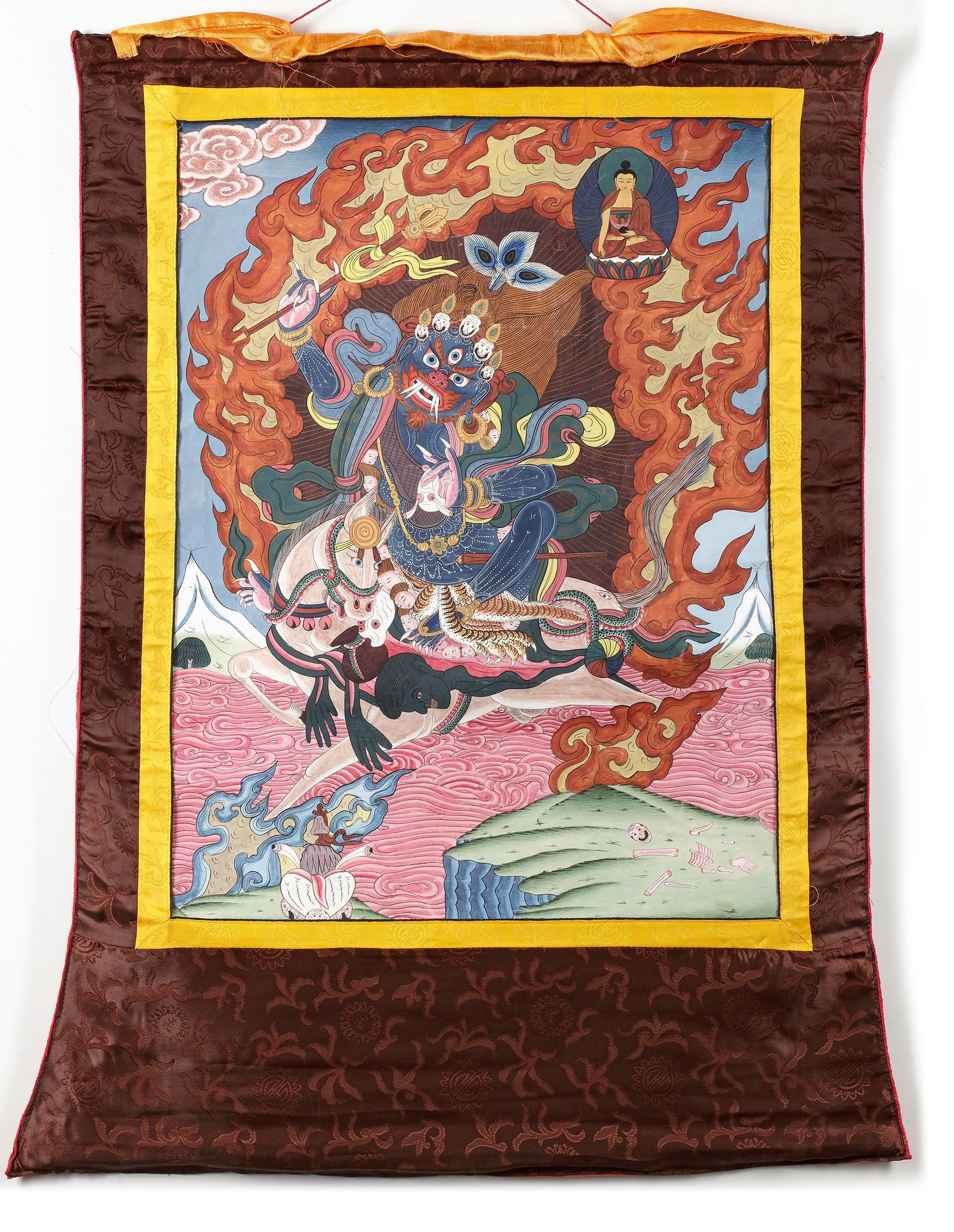 A WRATHFUL DEITY THANGKA, TIBET, 20TH CENTURY: A dramatic Tibetan thangka depicting a fierce wrathful Buddhist protector deity surrounded by flames, riding across a mystical landscape beneath a seated Buddha. Richly painted in vibrant mineral pigm
