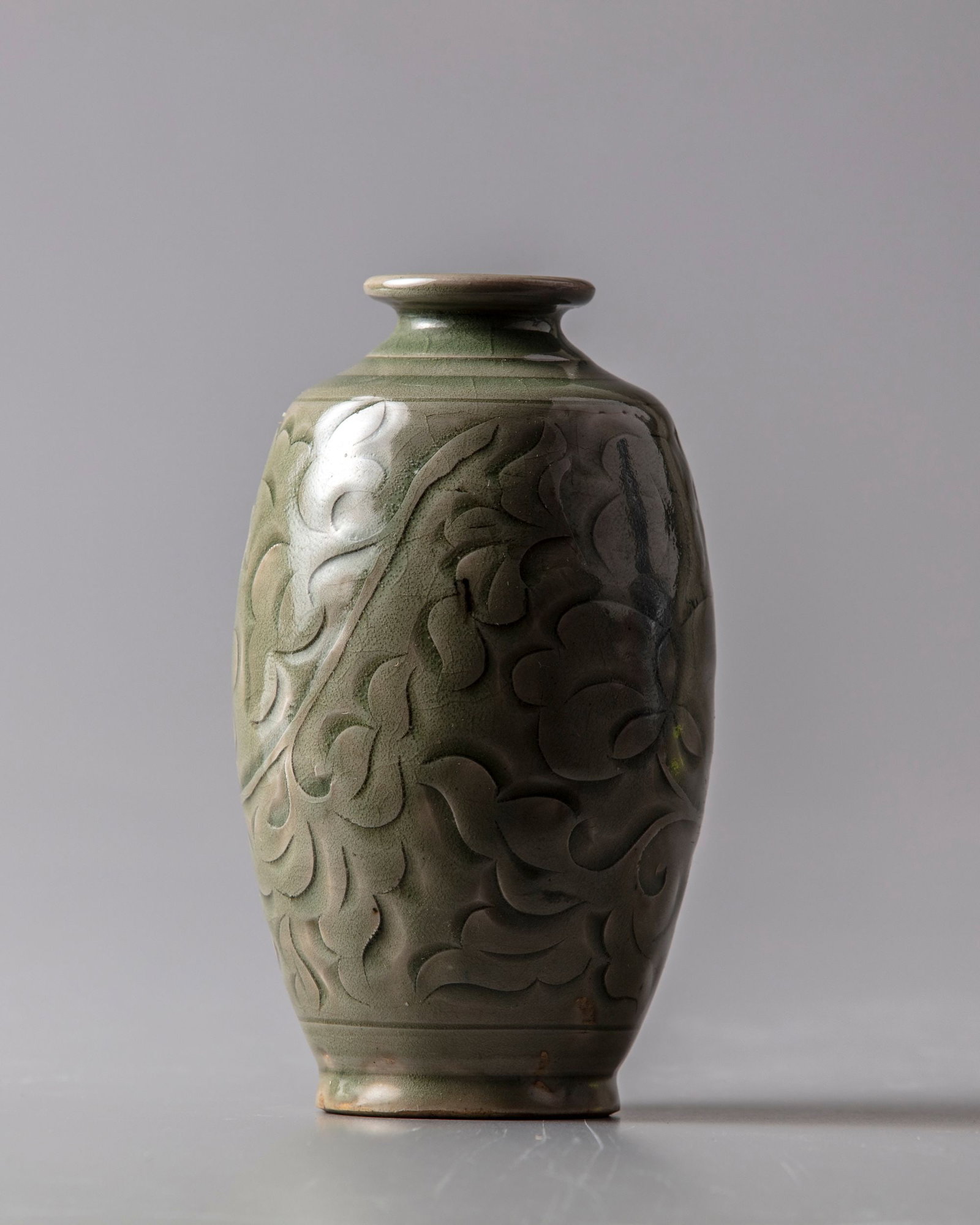 A CHINESE CELADON GLAZED VASE: With sloping shoulders and tapering sides below a short, straight neck, the vase is freely carved around the exterior with leafy chrysanthemum sprays. The whole is covered in a glaze of rich olive-gre