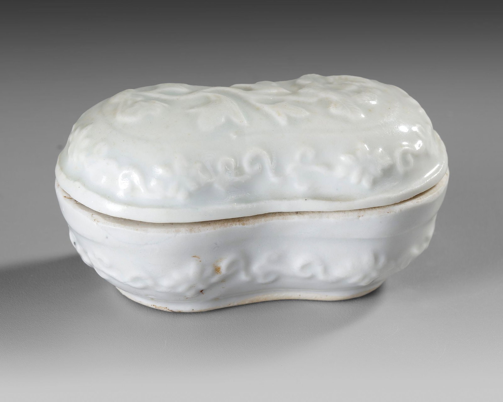A CHINESE WHITE-GLAZED COVERED BOX, 17TH CENTURY: Of compressed peanut-shaped form, the box is surmounted by a domed cover molded in high relief with a sinuous dragon emerging amidst scrolling clouds. The matching base is similarly decorated with a c
