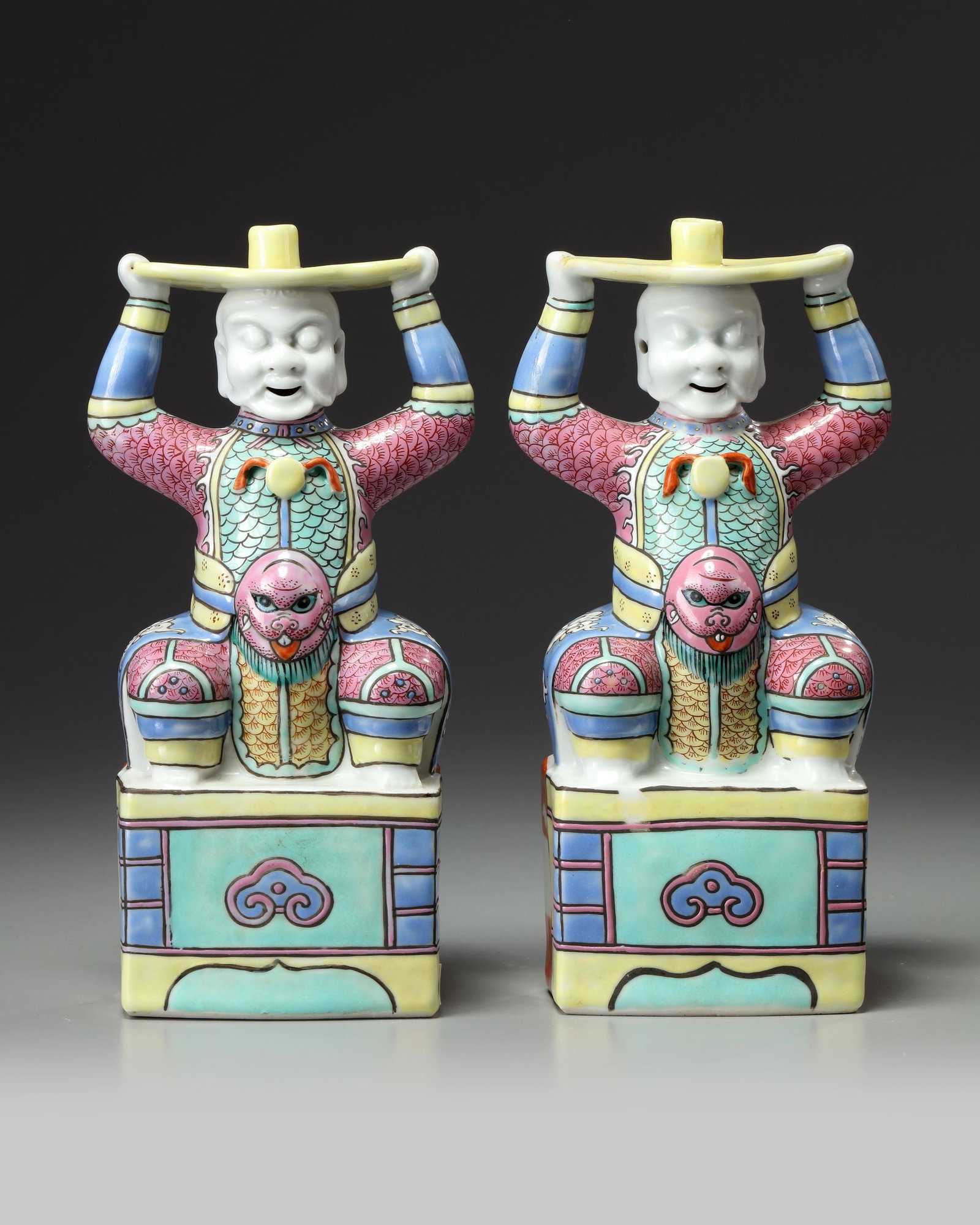A PAIR OF CHINESE FAMILLE ROSE FOREIGNER CANDLESTICKS, 19TH CENTURY: Each candlestick is modeled in the form of a bearded pot-bellied foreigner wearing armor, seated on a rectangular base, balancing the yellow-enamelled ingot-shaped drip plate upon his head, supported 
