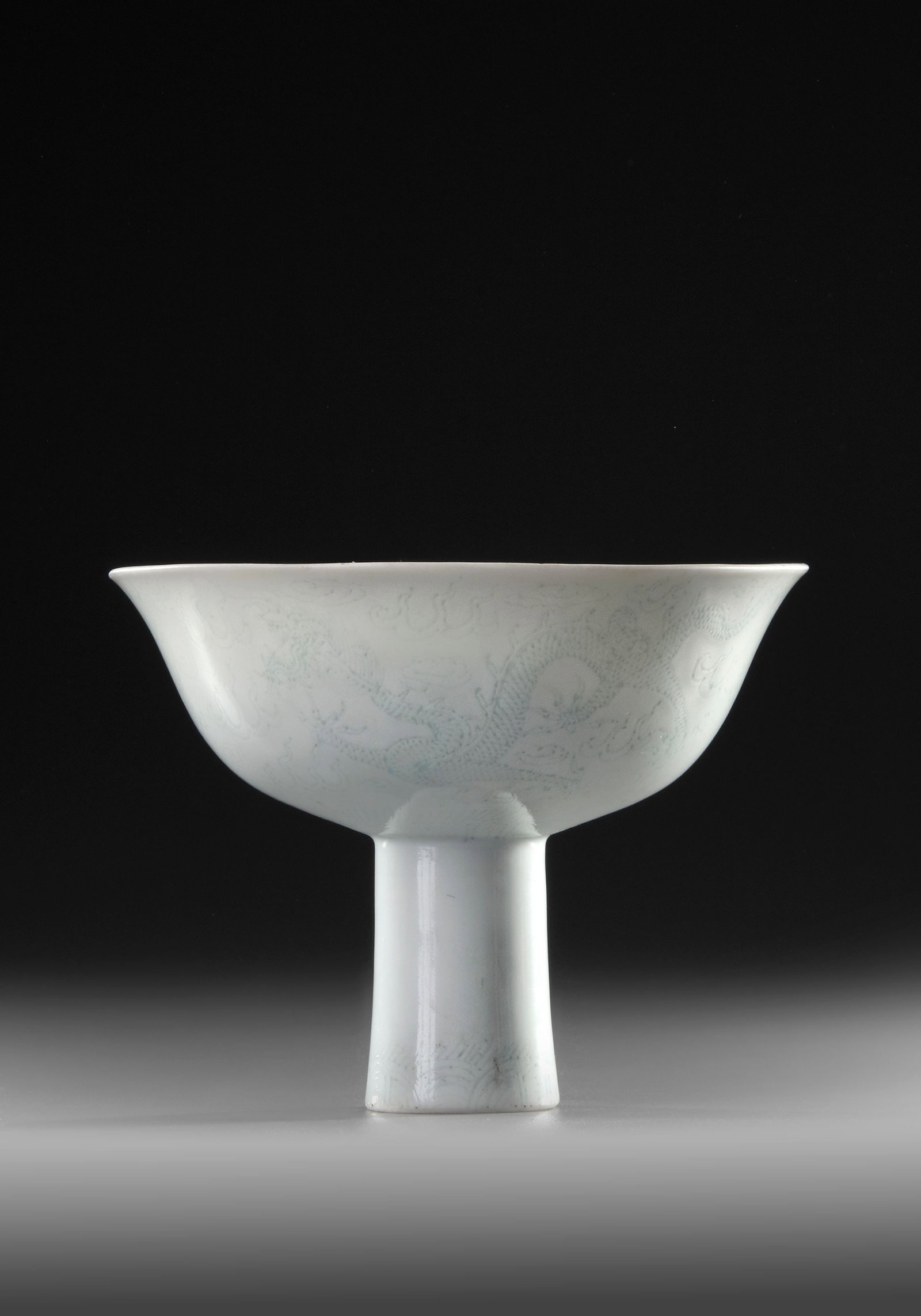 A CHINESE WHITE-GLAZED STEM BOWL, 17TH CENTURY: A Chinese porcelain stem cup standing on a small flaring foot with flaring sides, covered overall with a pale ice-blue translucent glaze, carved to the exterior and the interior with two five-clawed d