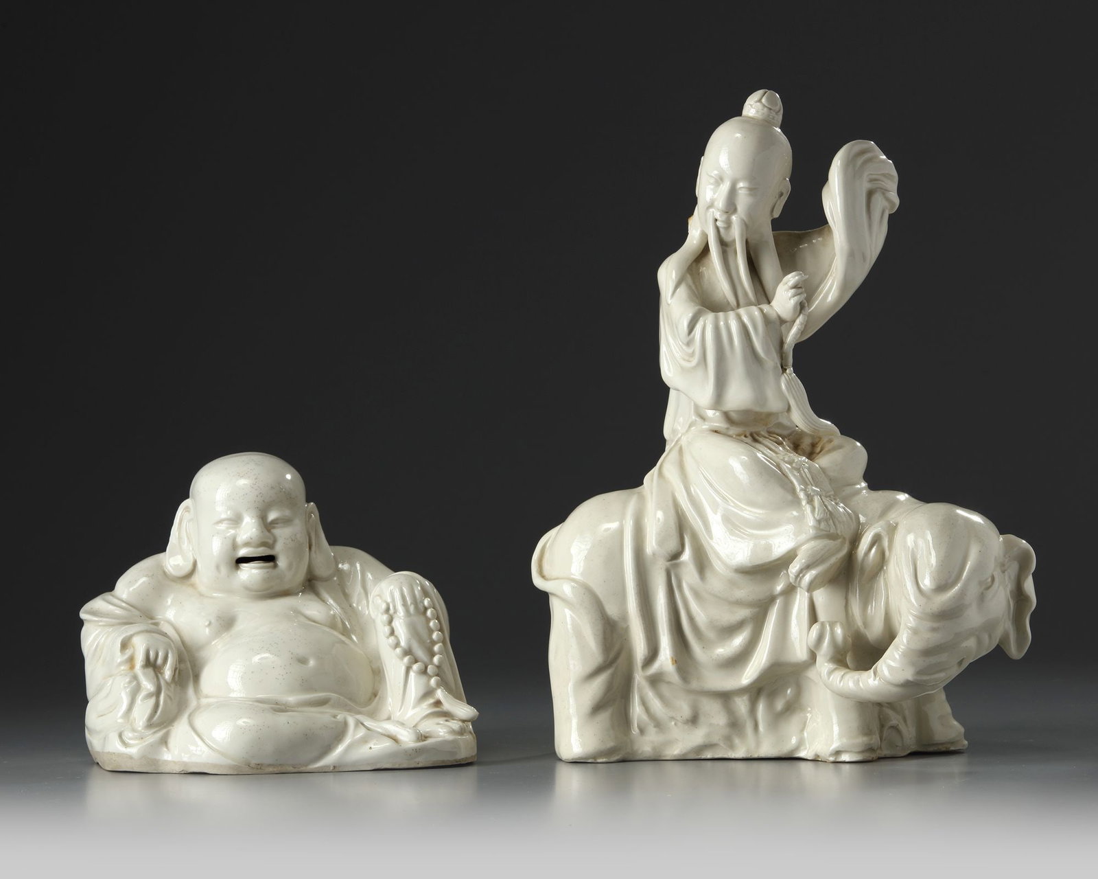 TWO CHINESE WHITE GLAZED IMMORTALS, 19TH CENTURY: Comprising a smiling Budai resting on his cloth bag, wearing long open robes with his left hand holding rosary beads, and a bearded Immortal riding on an elephant. Budai: 13.5 cm. high by 17.5 cm. wid