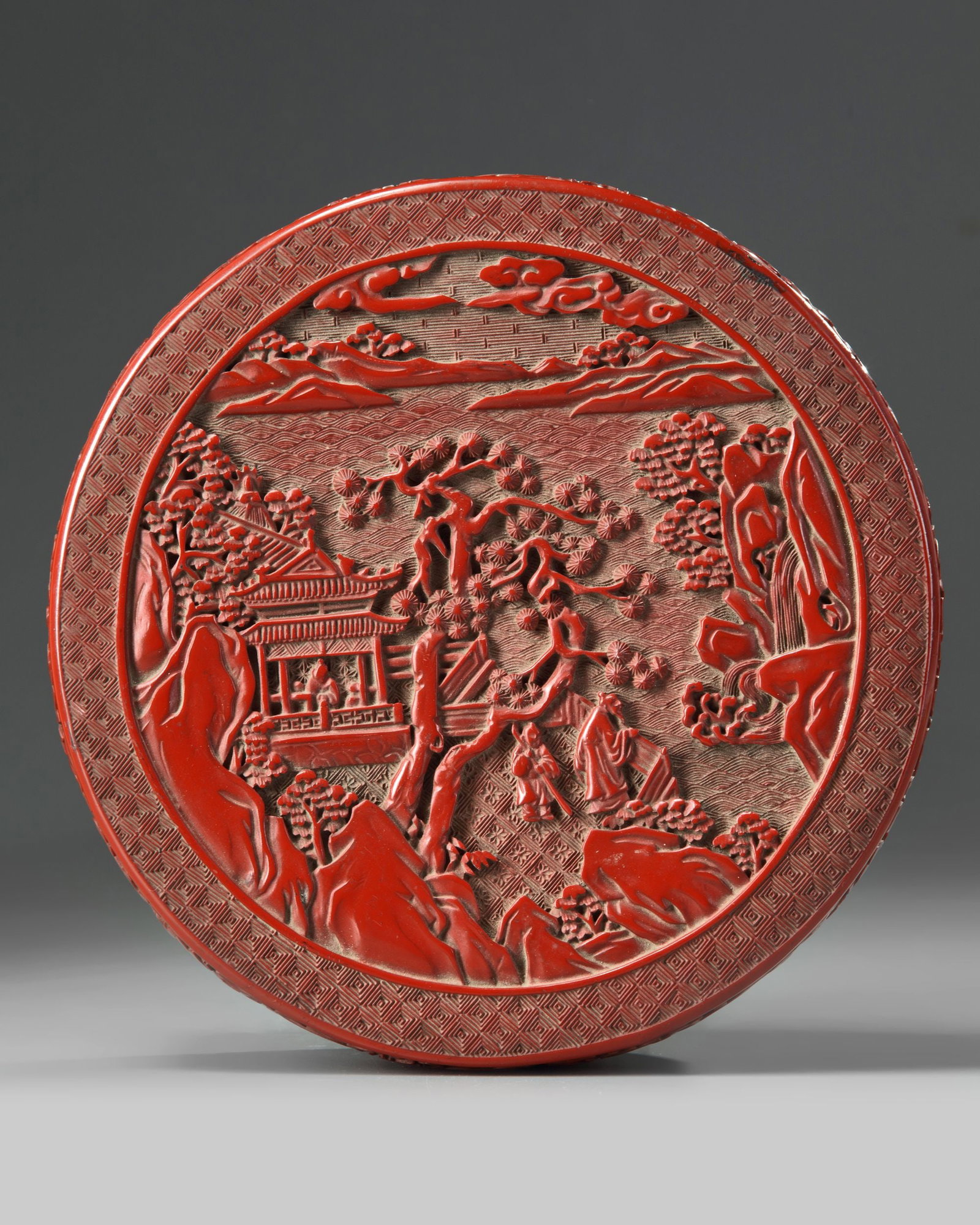 A CHINESE LACQUER BOX WITH A GARDEN SCENE, 19TH-20TH CENTURY: A carved red lacquer box and cover of rounded form, carved on the cover with attendants in a garden within a plain border. The sides are carved with lotus scrolls. 22 by 8 cm. Tiny surface scratches t