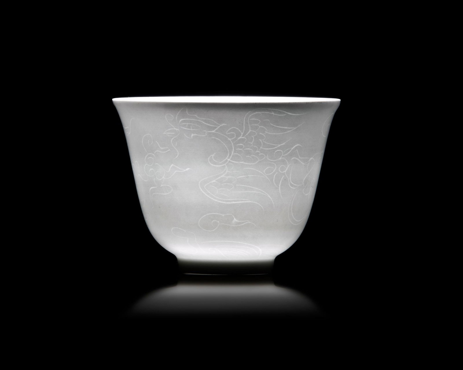 A SMALL CHINESE WHITE-GLAZED DRAGON AND PHOENIX CUP, 20TH CENTURY: The cup is decorated with two striding anhua dragon and phoenix above tumultuous waves. The base bears an apocryphal Tian mark. Height: 5 cm. Diameter: 6.7 cm. Appears to be in a good condition.