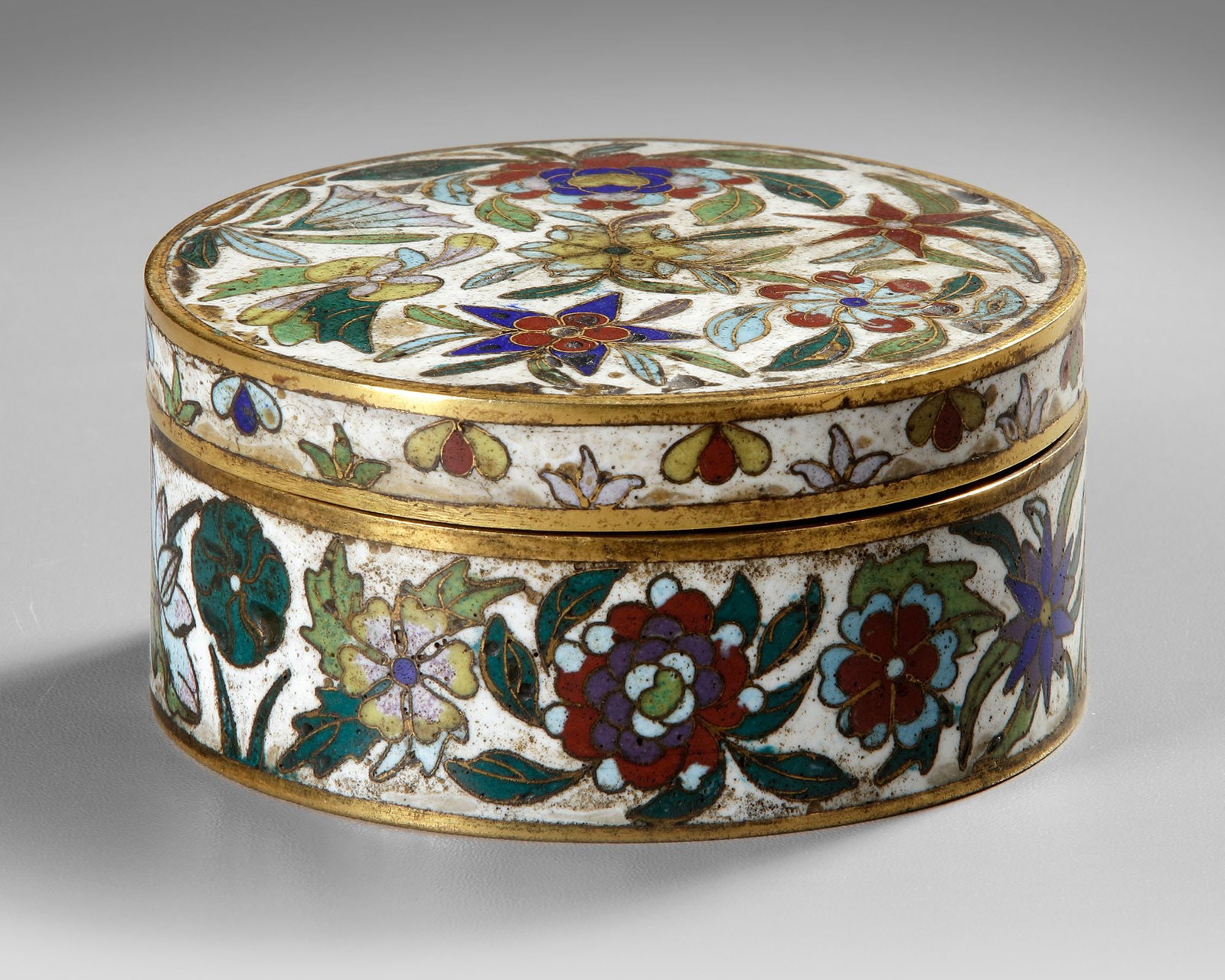 A CHINESE CLOISONN?‰ ENAMEL BOX, 19TH CENTURY: Of circular section, the cover and the sides brightly enameled with lotus, lilies and peonies blossoming branches on a white ground. Height 4.5 cm. Diameter 9.3 cm.