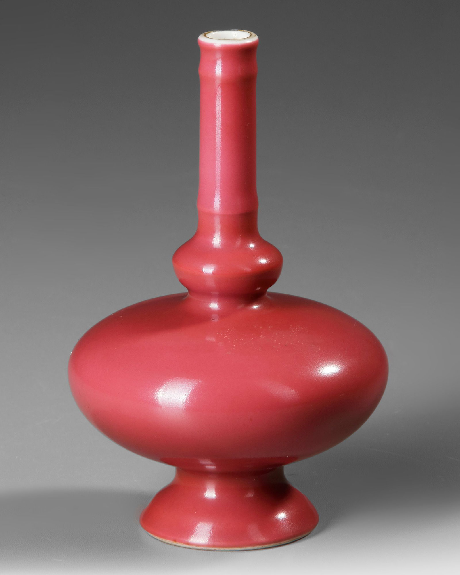 A SMALL CHINESE RUBY-GLAZED VASE, 19TH-20TH CENTURY: A Chinese porcelain vase, the compressed globular body rising from a spreading foot to a tall, slender cylindrical neck with a raised rib encircling its midsection, subtly evoking a bamboo form. The v