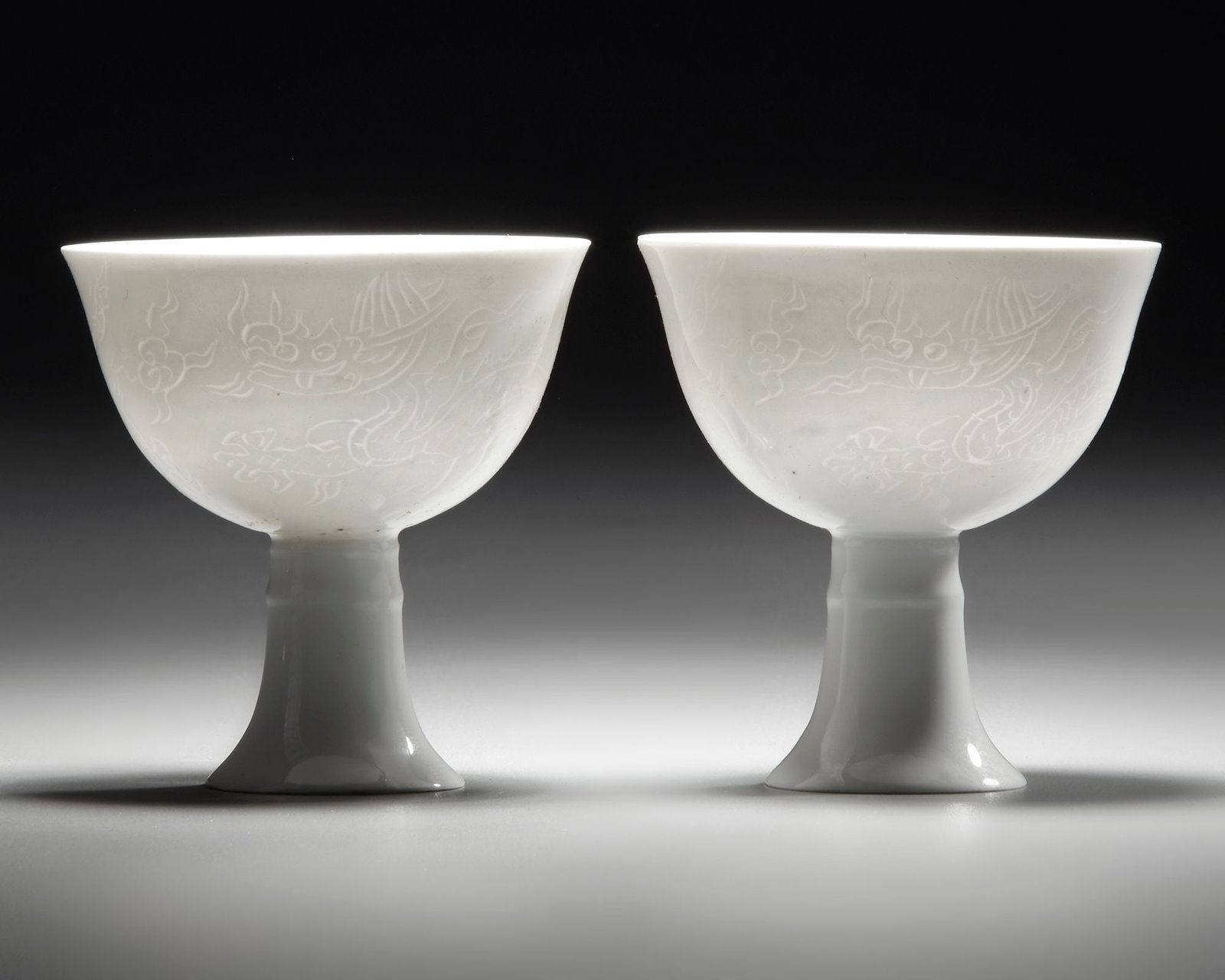 A PAIR OF CHINESE WHITE-GLAZED DRAGONS STEM CUPS, 20TH CENTURY: Each stem cup is decorated with two striding anhua dragons above tumultuous waves. The base bears an apocryphal Chenghua mark. Diameter: 7.8 cm. Height: 8 cm.