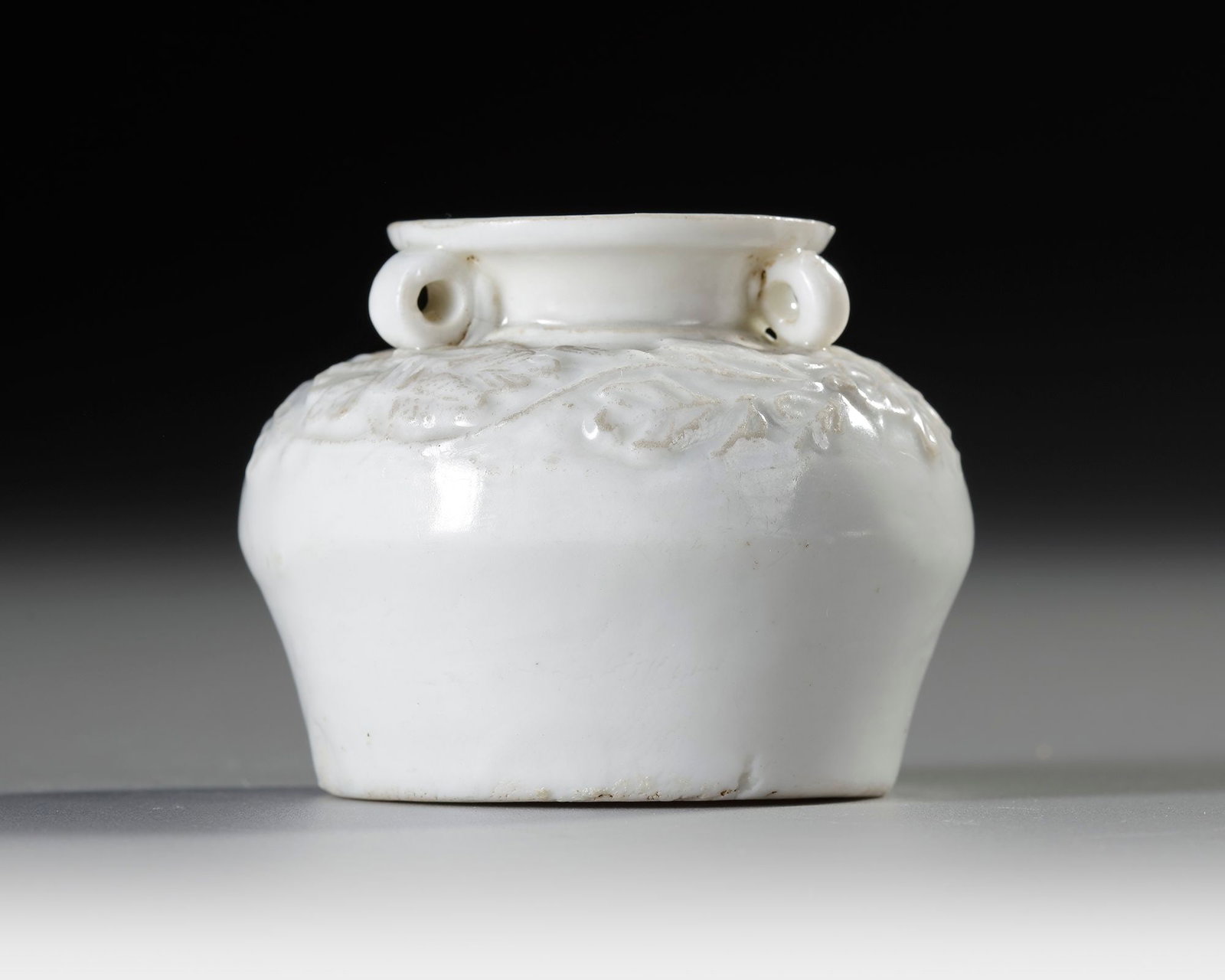 A CHINESE DEHUA GLAZED MINIATURE VASE, MING DYNASTY (1368-1644): A Chinese Dehua porcelain miniature vase, of compressed globular form with short flared neck and opposing loop handles, the shoulder molded in relief with scrolling floral decoration beneath a creamy 