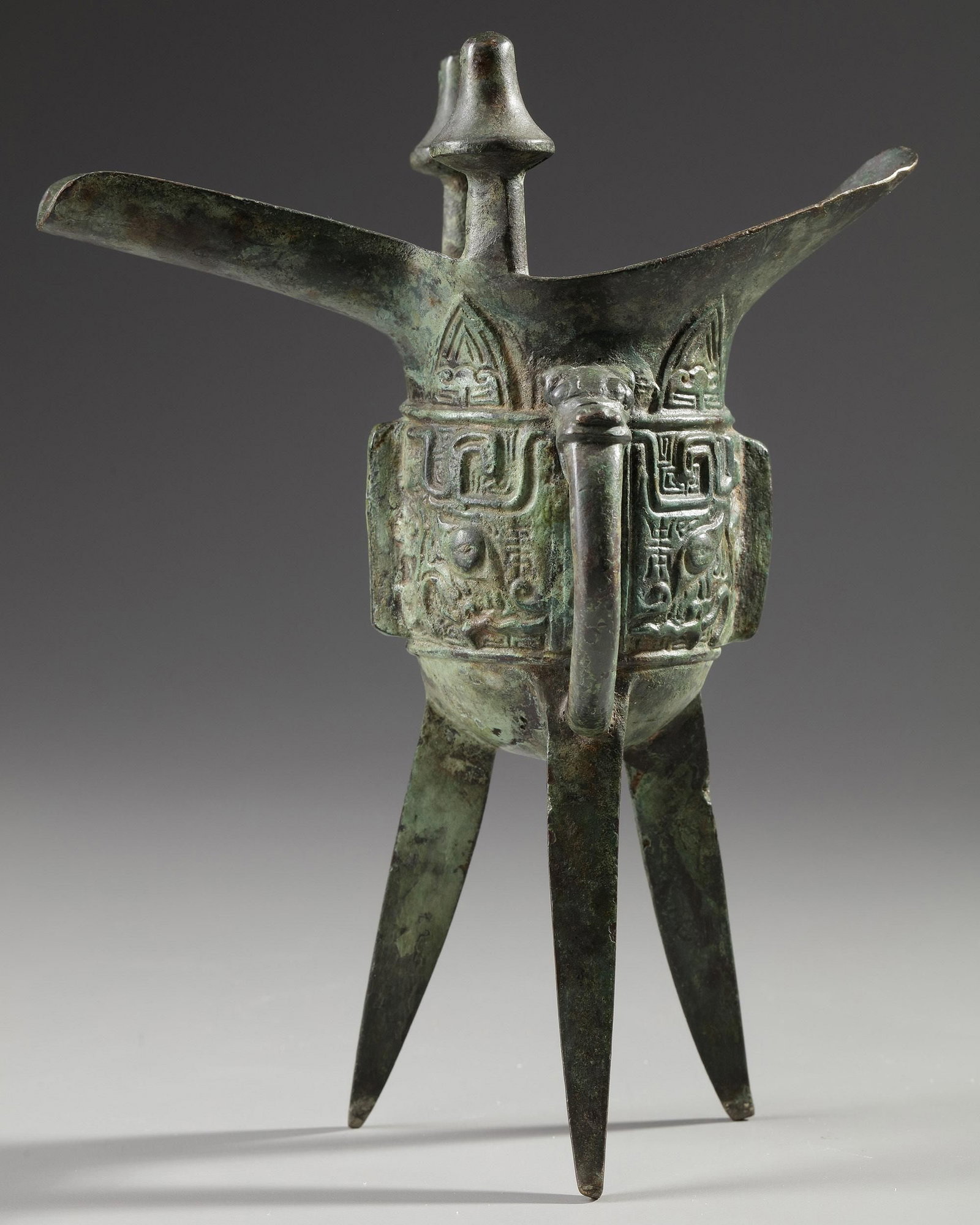 A CHINESE BRONZE JUE, QING DYNASTY (1644-1911): Raised on three blade-form supports, the sides cast with a band of two taotie masks reserved on a leiwen ground and separated by narrow notched flanges, one mask centered on a third flange, the other 