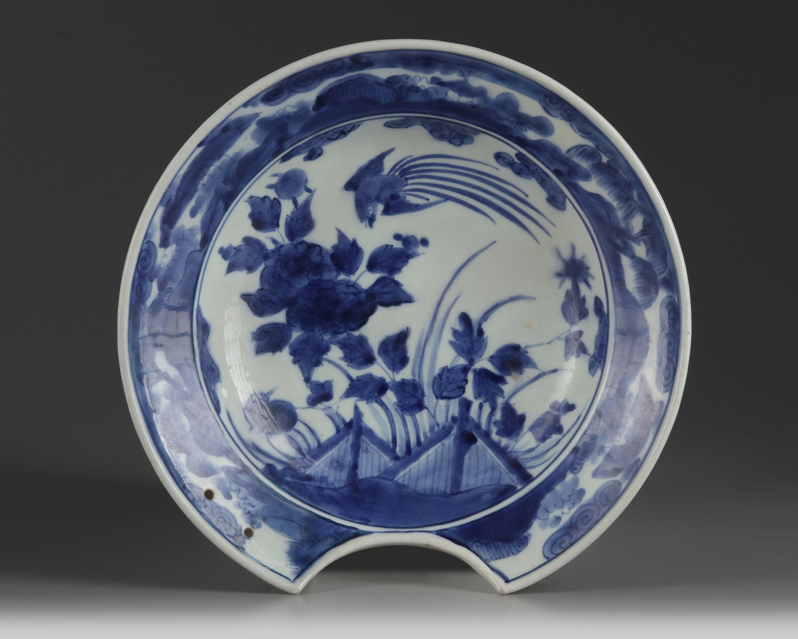 A JAPANESE ARITA SHAVING BOWL, 18TH CENTURY: Shaving bowl or barbers bowl on footring, spreading flat rim with a semicircular section saved in the lower part, with two pierced. Decorated in underglaze blue with a bird in flight above peonies in 