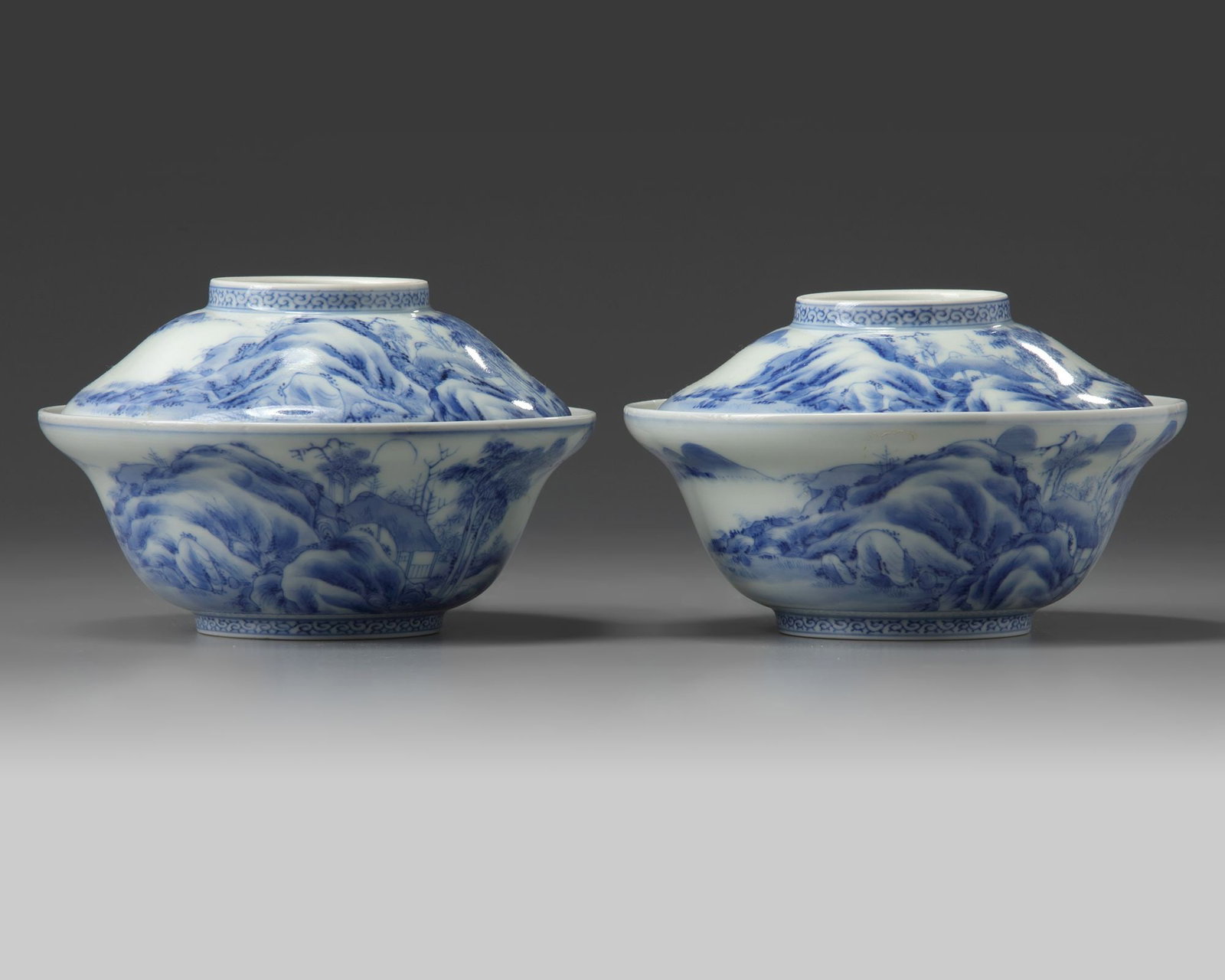 A PAIR OF JAPANESE HIRADO BOWLS AND COVERS, 19TH CENTURY: Standing on a small footring, decorated in underglaze blue with houses amongst trees in a mountainous landscape. Height 10 cm. Diameter 15 cm. A tiny chip to the rim of one bowl.