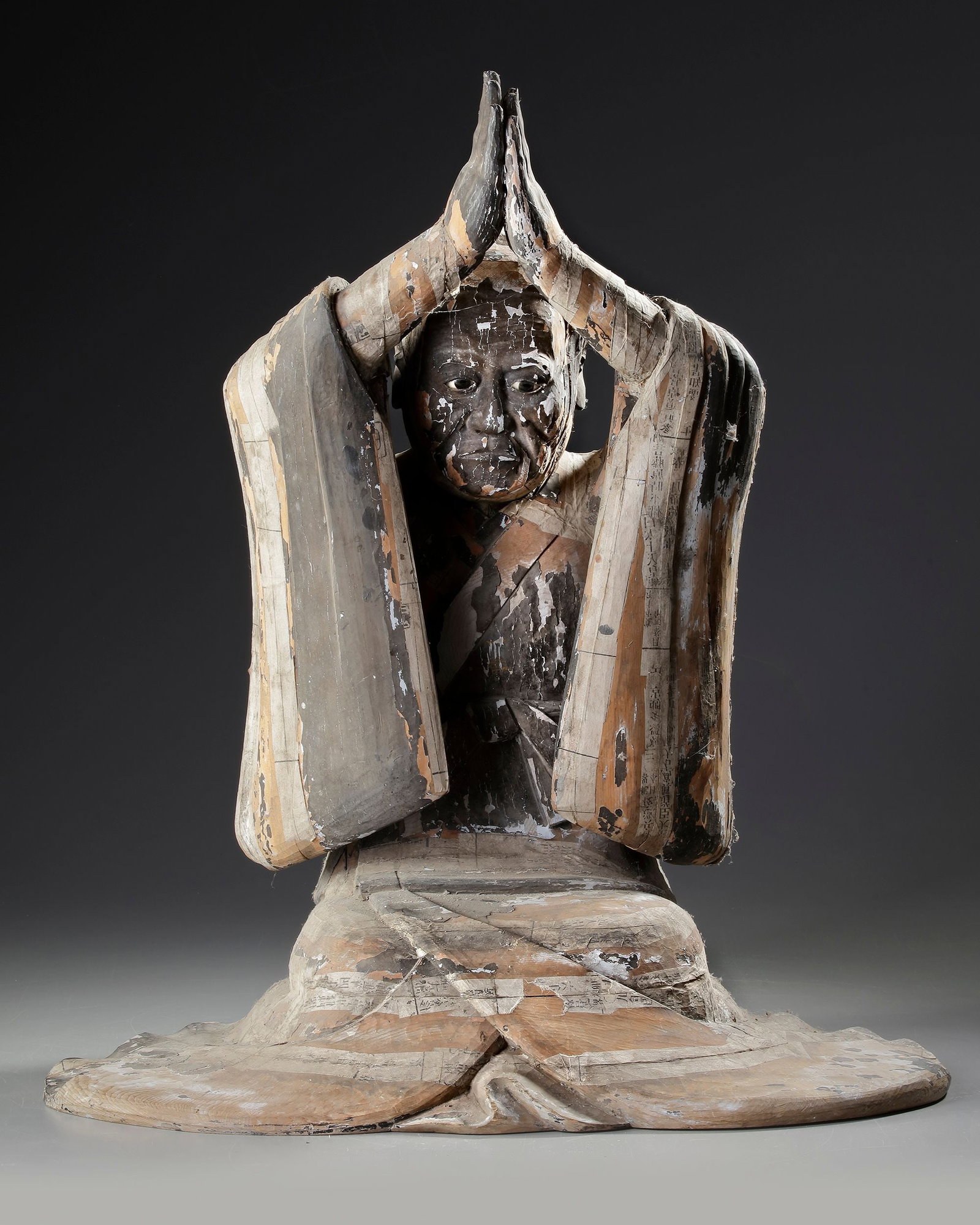 A JAPANESE WOODED FIGURE OF A DEVOTEE IN PRAYER, 16TH CENTURY: Carved as a kneeling figure with hands raised above the head and pressed together in an attitude of devotion, the sculpture is portrayed wearing voluminous monastic robes that fall in broad, angular f