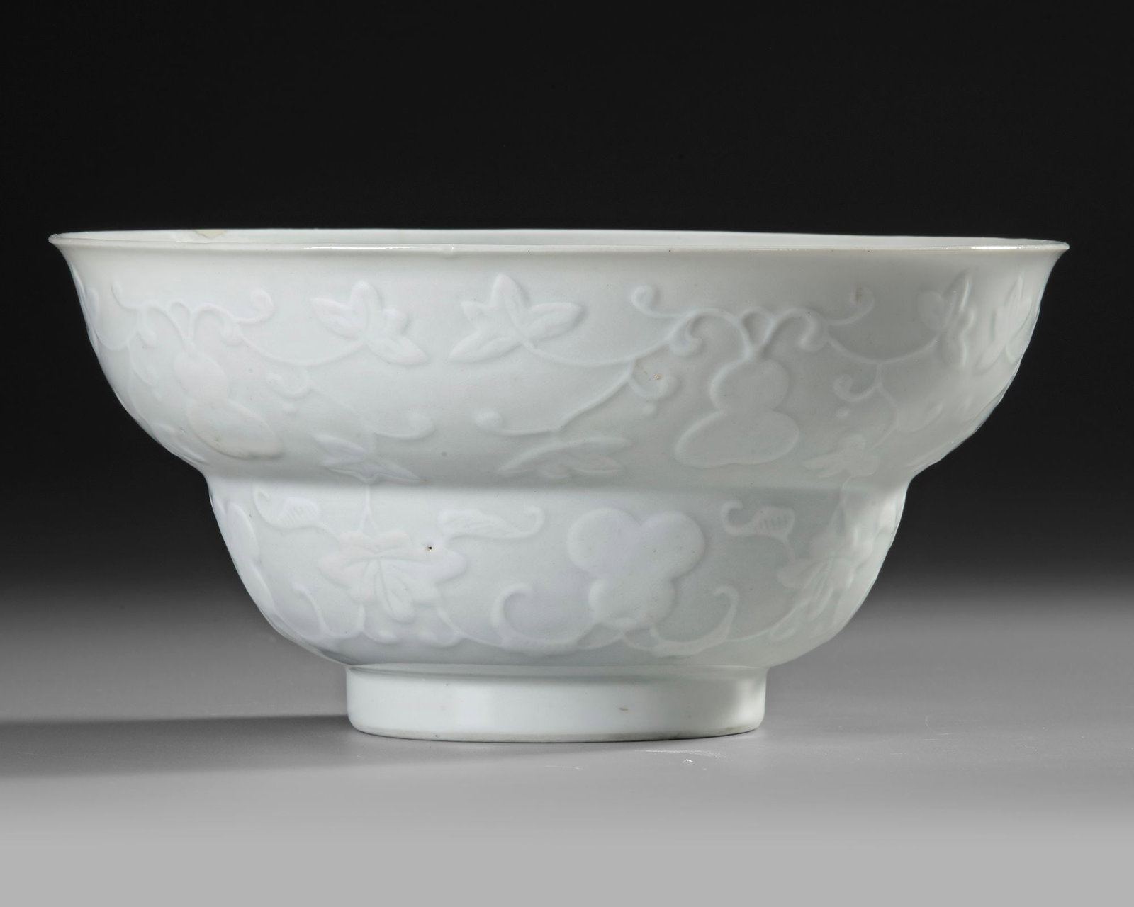 A CHINESE WHITE GLAZED PORCELAIN STEMMED BOWL, KANGXI PERIOD: A Chinese porcelain stemmed bowl, finely moulded in relief with scrolling flowering and fruiting branches, the rounded body raised on a short spreading foot. The elegant monochrome white decoration is