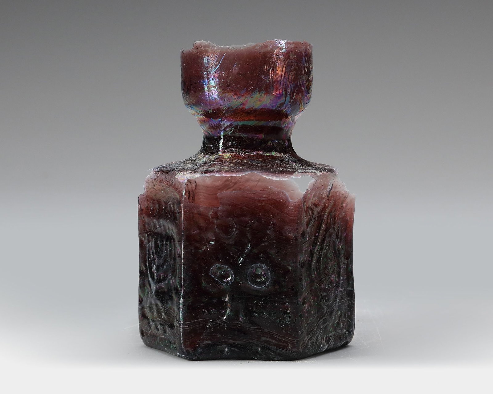 A SIDONIAN MOLD-BLOWN BOTTLE SIDON, 1ST CENTURY AD: Mold-blown glass bottle with a hexagonal (six-sided) body and a short cylindrical neck, produced in dark reddish-purple glass. The flat panels are decorated with molded geometric and scroll motifs. He