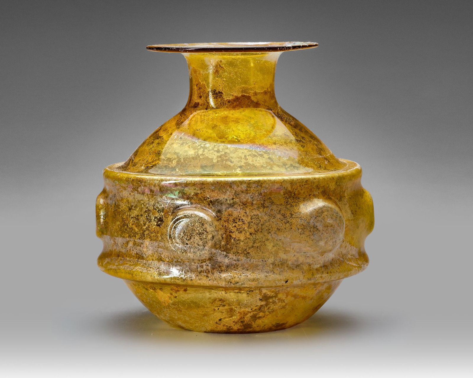 AN AMBER GLASS VESSEL, PERSIA OR SYRIA, 11THâ€“12TH CENTURY: Free-blown amber glass vessel with a compressed globular body and short, flaring neck. The body is encircled by a raised applied band with rounded bosses, providing both decoration and grip. Height 9.