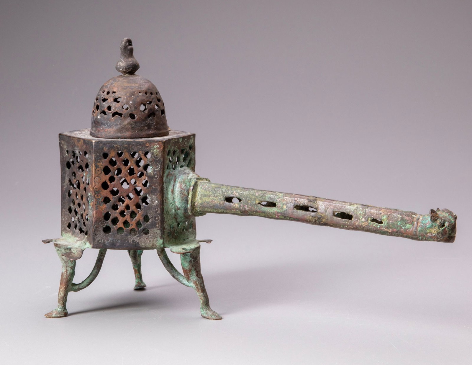 A KHORASAN BRONZE INCENSE BURNER, PERSIA, 12TH CENTURY: Cast bronze, of rectangular form on four slender feet, the sides extensively pierced with geometric openwork, surmounted by a domed, pierced cover with a finial. The body fitted with a long tubular, p