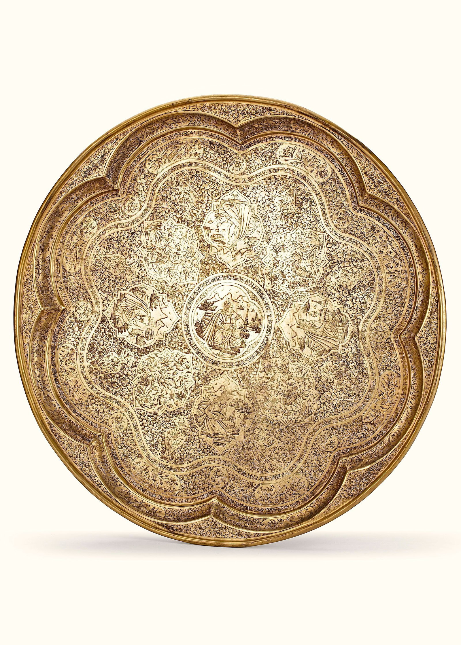 A QAJAR BRASS ENGRAVED TRAY, PERSIA, 19TH CENTURY: A large circular brass tray with a lobed rim, finely engraved with dense floral motifs and figural scenes. The central medallion features a finely detailed scene of a seated figure smoking a hookah in