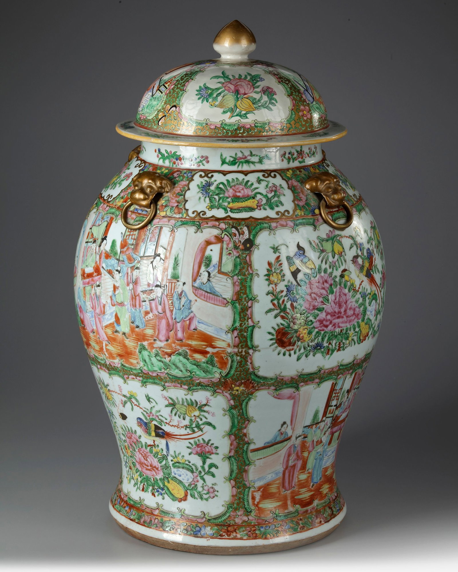 A CHINESE FAMILLE ROSE CANTON VASE WITH COVER, 19TH CENTURY: A Chinese porcelain vase stylised with four gilt Fo lions heads handles with rings, enameled with panels of court scenes and birds perched amongst peonies blossoming branches, divided by flower scroll