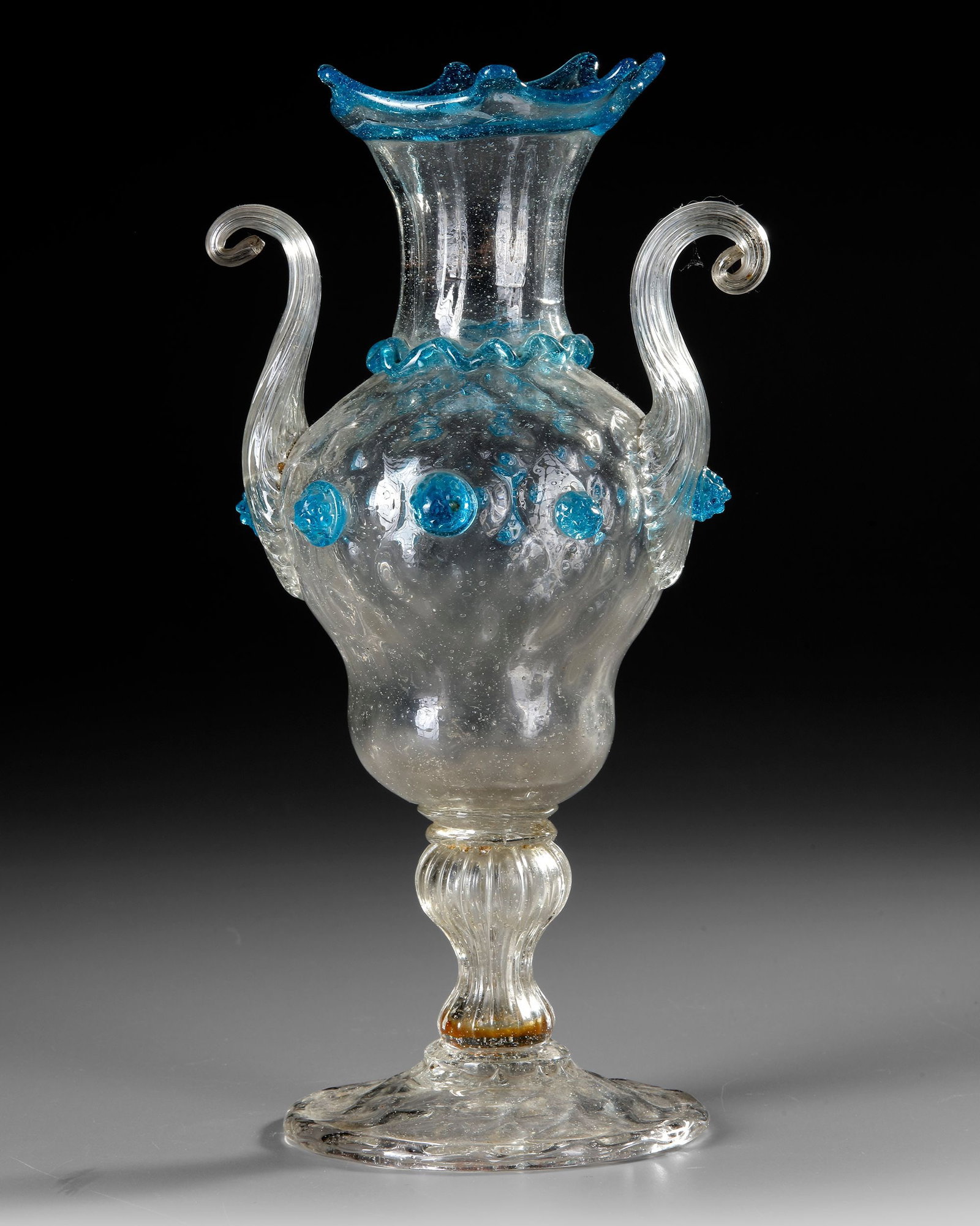 ITALIAN GLASS VASE VENICE, EARLY 17TH CENTURY: An elegant Venetian glass vase distinguished by its clear, finely blown body, bright blue applied decoration, and graceful scrolling handles. The delicate foot, elongated neck, and crown-like blue rim