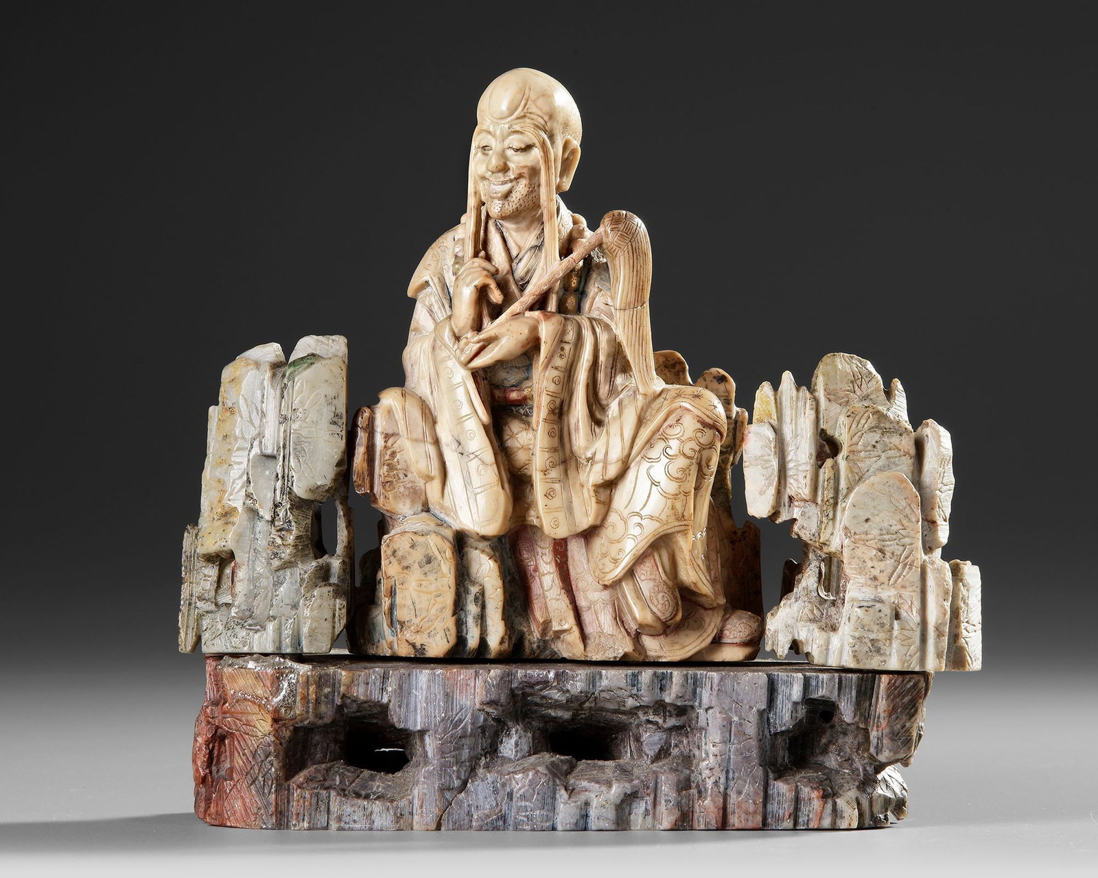 A CHINESE SOAPSTONE FIGURE OF A DAOIST IMMORTAL, 19TH CENTURY: Finely carved in variegated soapstone, depicting a Daoist Immortal seated amidst a dramatically sculpted rocky landscape. The figure is shown with a serene, smiling expression, elongated earlobes and 