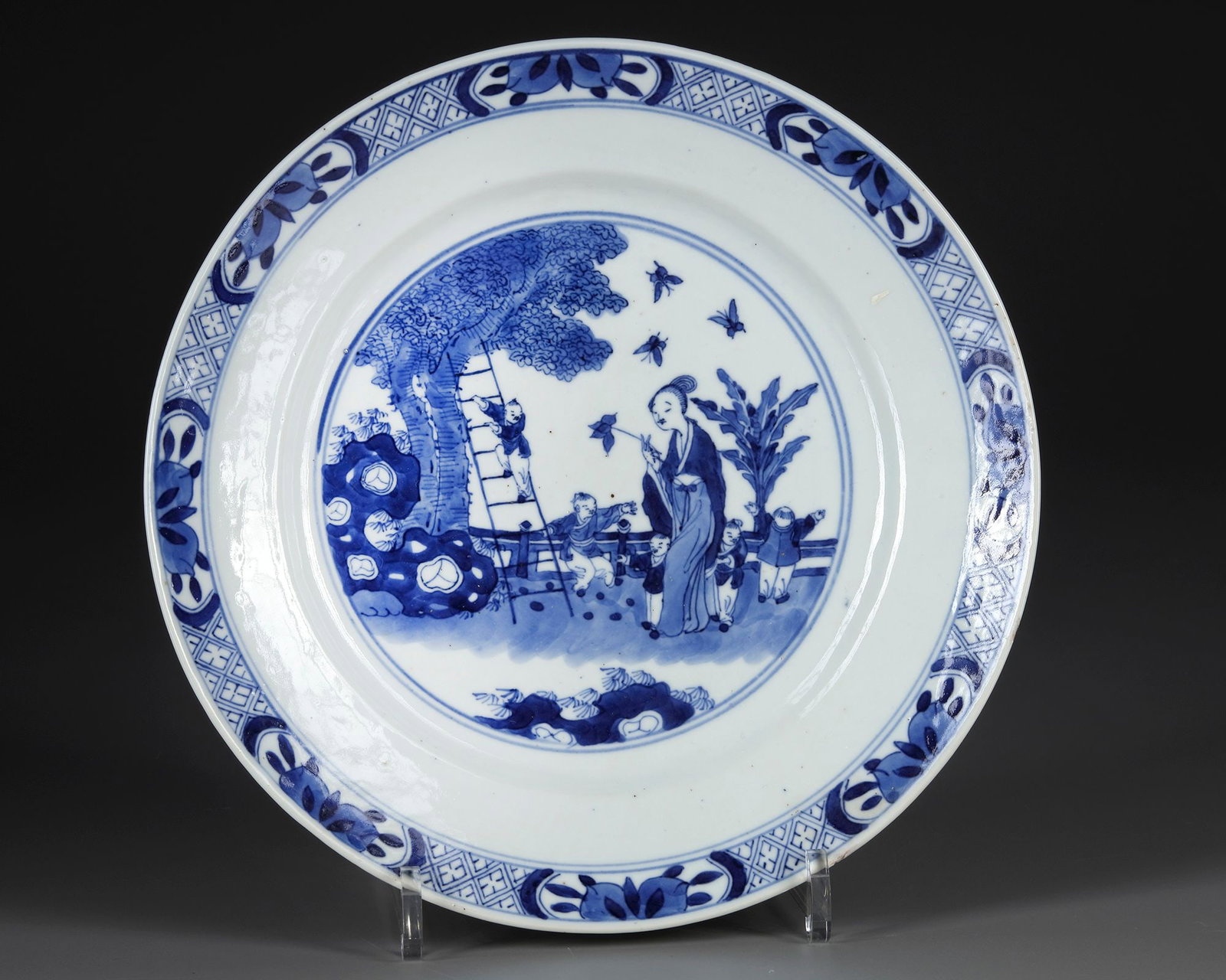 A CHINESE BLUE AND WHITE PORCELAIN PLATE WITH A KANGXI MARK, 19TH CENTURY: Chinese blue and white porcelain plate, the central roundel painted with a continuous garden scene depicting a lady accompanied by children beneath a spreading tree, with butterflies and ornamental ro
