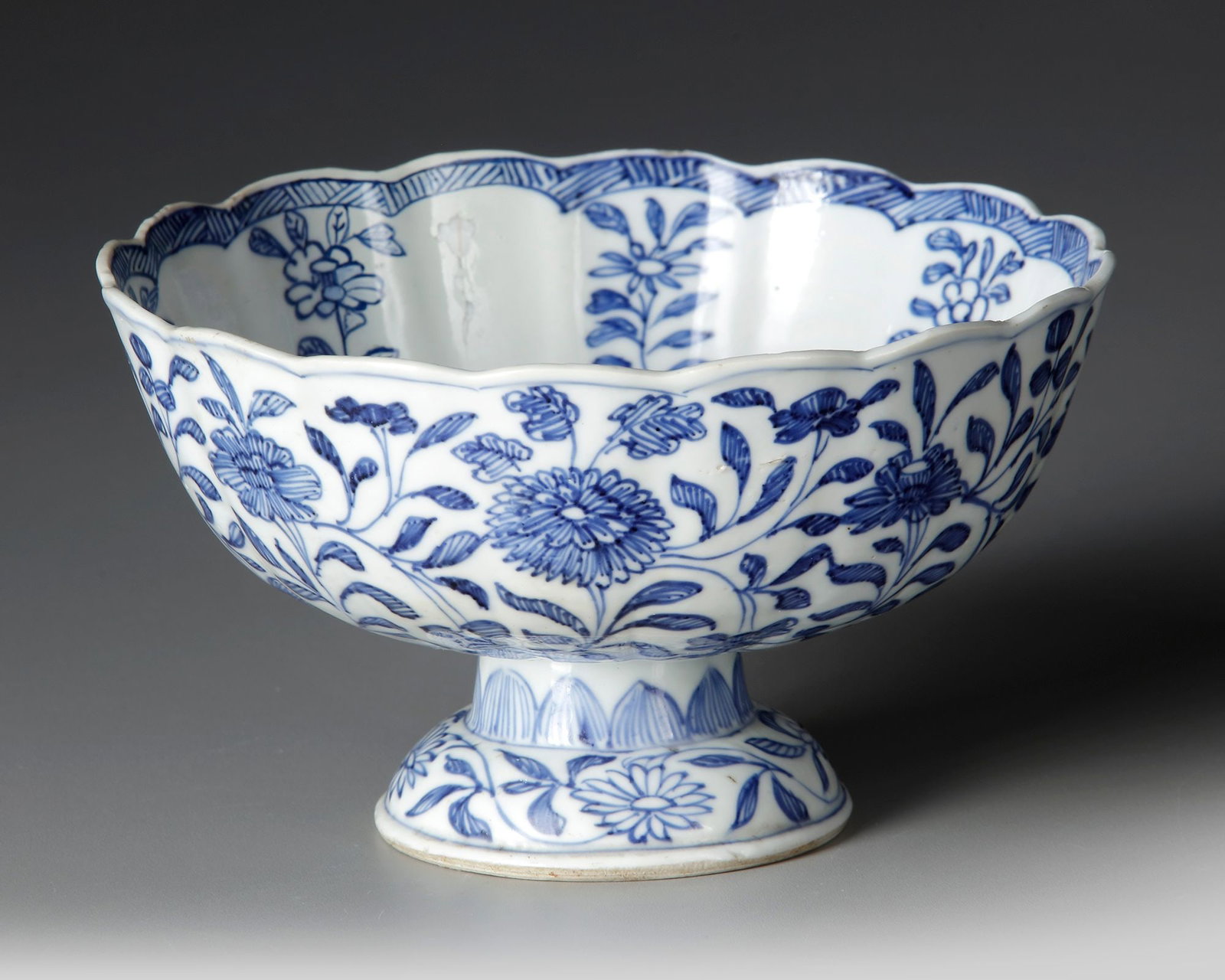 A CHINESE BLUE AND WHITE PEDESTAL BOWL, KANGXI PERIOD,(1662â€“1722): A Chinese blue and white porcelain pedestal bowl, the broad rounded bowl rising from a waisted stem and spreading foot, the scalloped rim decorated with a narrow diaper border. The exterior is finely 