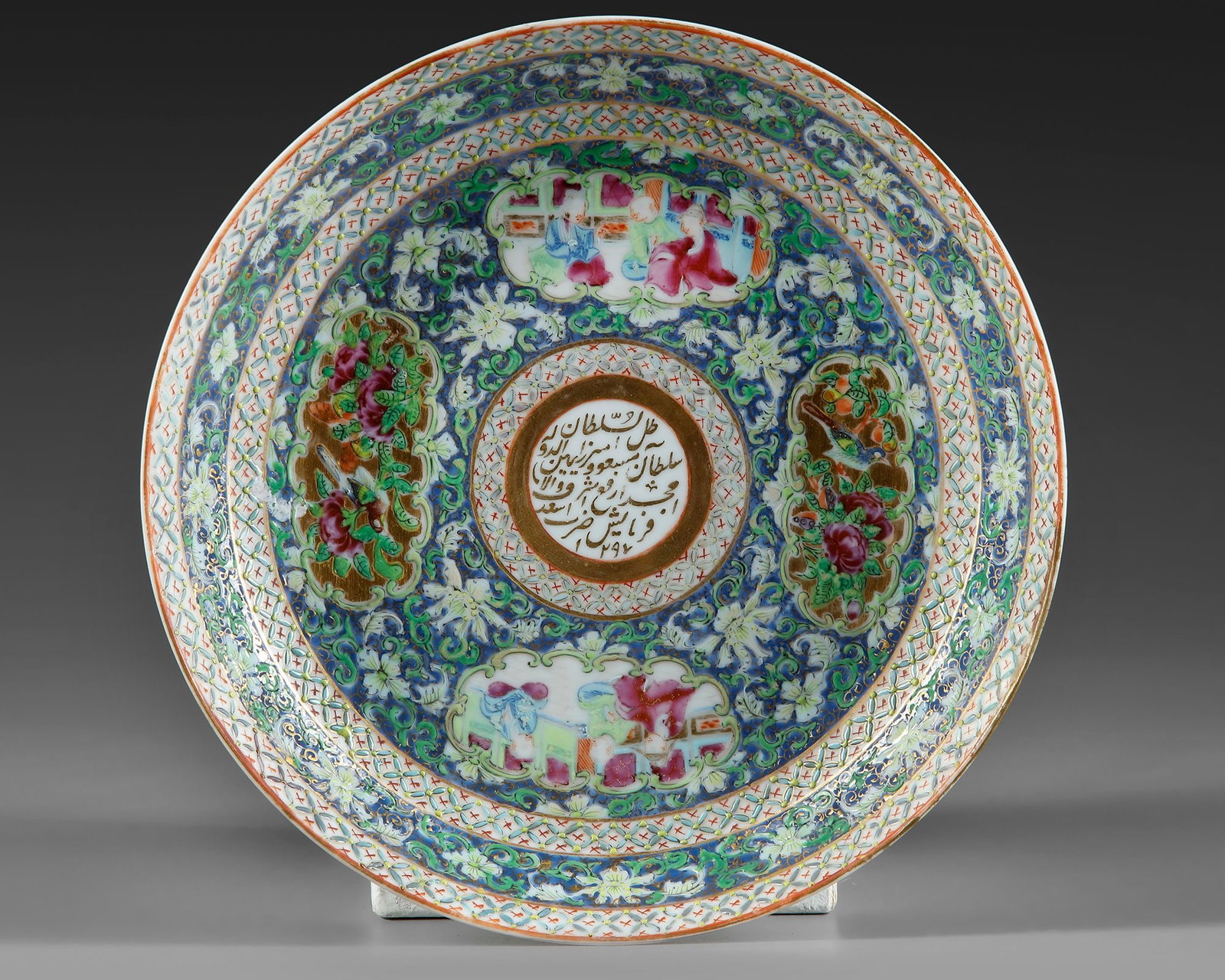 A CANTONESE EXPORT PORCELAIN DISH MADE FOR ZILL AL-SULTAN, CHINA, DATED 1297 AH/1879-80 AD: The dishe of shallow rounded form, extensively enamelled in polychrome with lotus sprays on a blue ground, with cartouches depicting figural scenes alternating with cartouches depicting floral sprays 