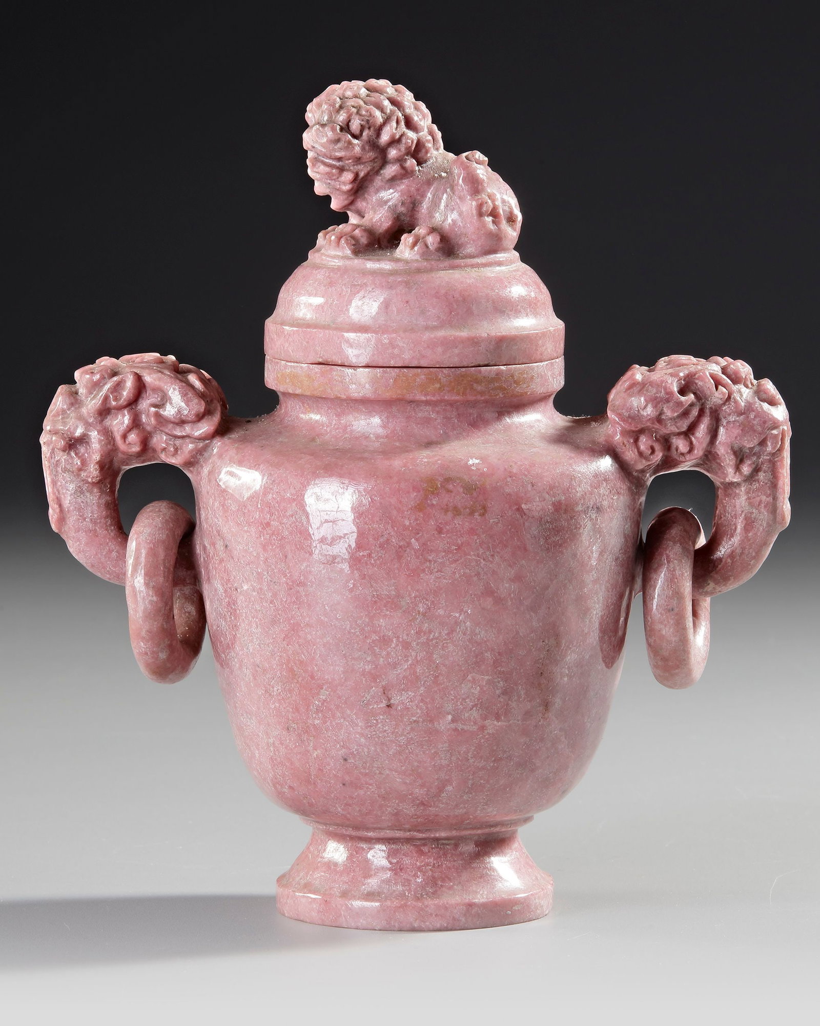 A CARVED PINK STONE LION VASE, CHINA, 20TH CENTURY: Decorative carved pink stone vase of baluster form, featuring lion-mask handles with loose rings and a finely carved guardian lion (foo dog) surmounting the lid. Executed in mottled rose-colored hards