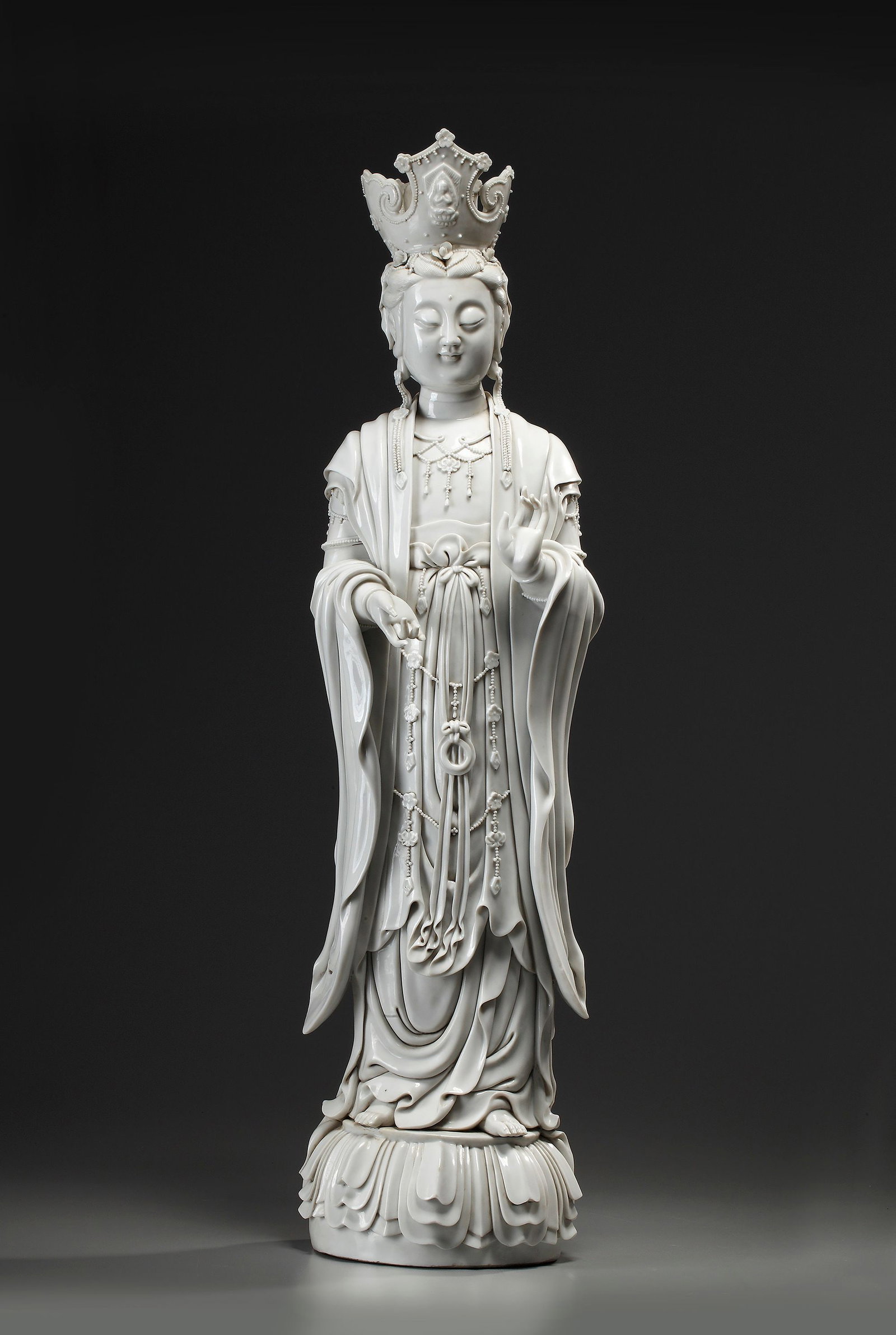 A LARGE CHINESE BLANC DE CHINE FIGURE OF GUANYIN, 19TH-20TH CENTURY: Standing on stylized lotus shaped base made up of multiple petals, dressed in an elaborate long swaying robe and richly decorated at the waist and chest with long strings of Buddhist jewellery with fl