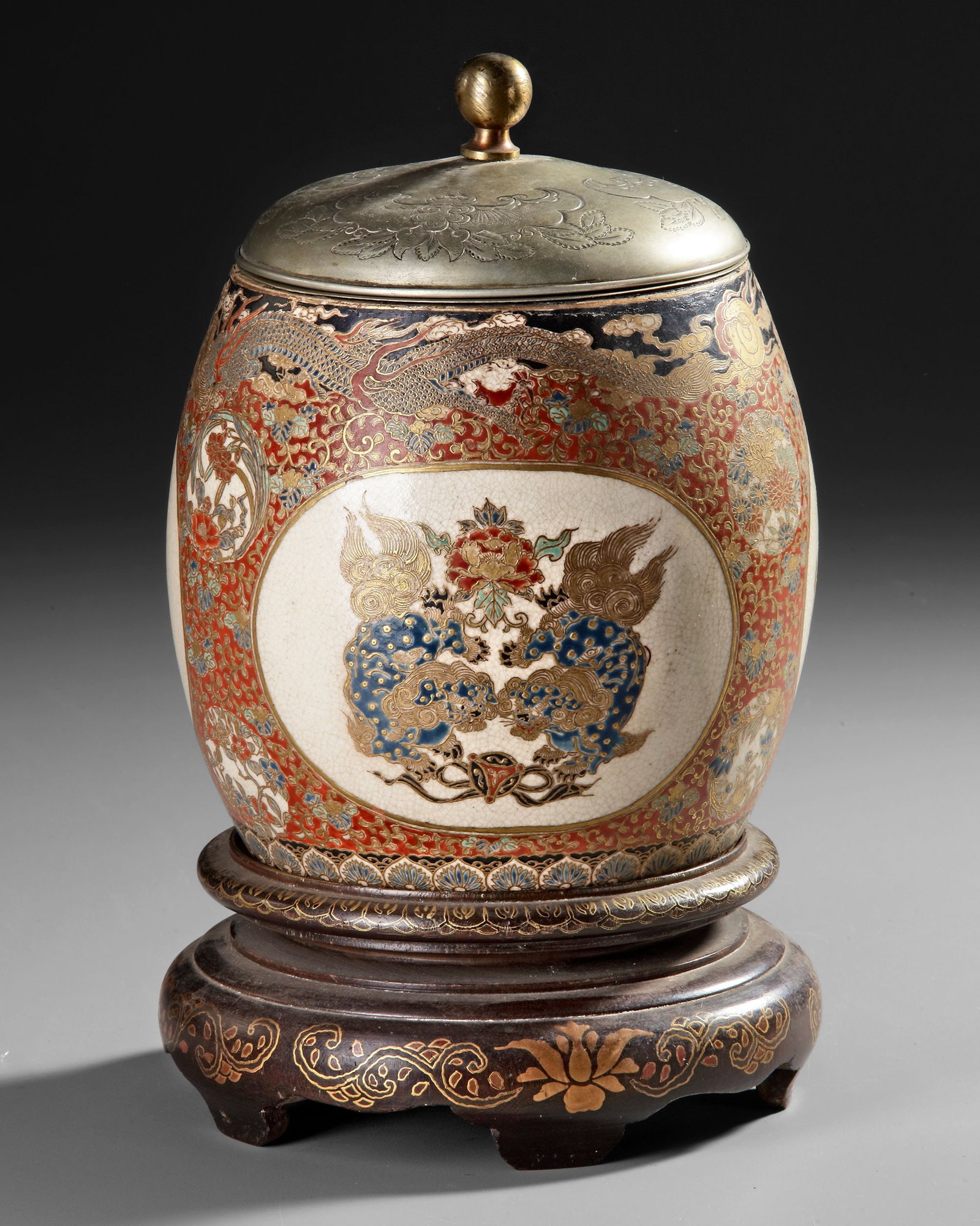 A JAPANESE SATSUMA COVERED JAR WITH WOODEN STAND, 19TH CENTURY: Finely decorated Japanese Satsuma earthenware jar with cover, richly embellished in polychrome enamels and gilt. The body features elaborate panels depicting mythical creatures, phoenix birds, and flo