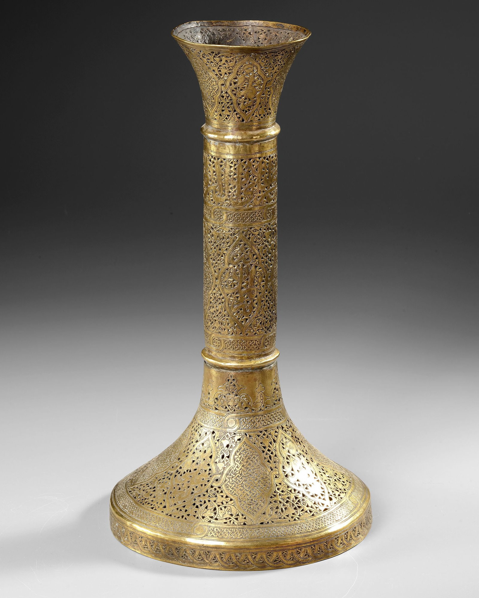 A QAJAR PIERCED BRASS CANDLESTICK, PERSIA, 19TH CENTURY: A Qajar brass candlestick of architectural form, featuring a broad, circular, stepped base, a tall cylindrical shaft and a flaring, trumpet-shaped candle nozzle. The entire surface is elaborately deco
