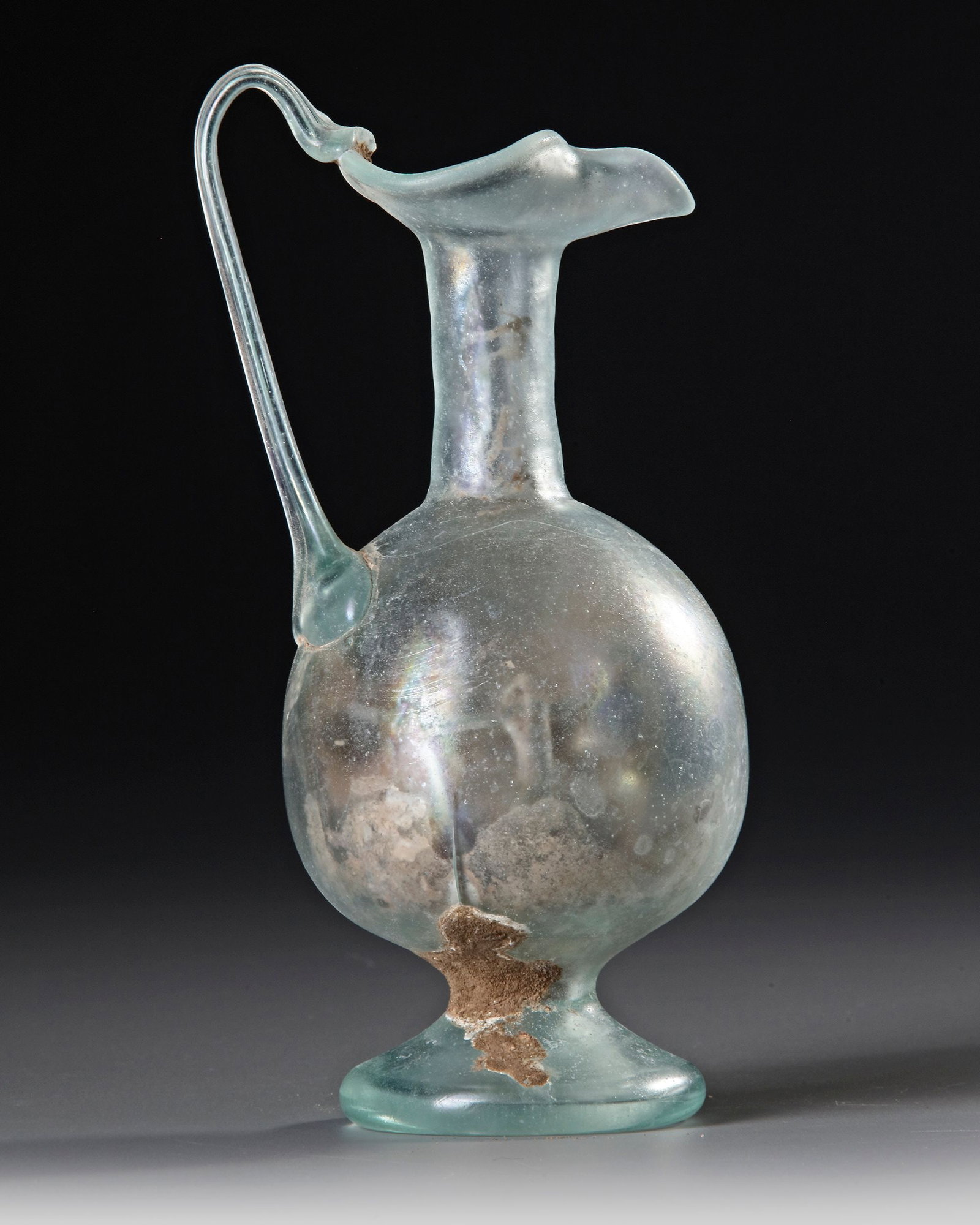 A ROMAN PALE GREEN GLASS EWER, CIRCA 1STâ€“3RD CENTURY A.D.: A fine pale green Roman glass ewer, with a rounded globular body, tall cylindrical neck and broad, elegantly flared rim. The vessel is distinguished by a slender applied handle rising in a graceful lo