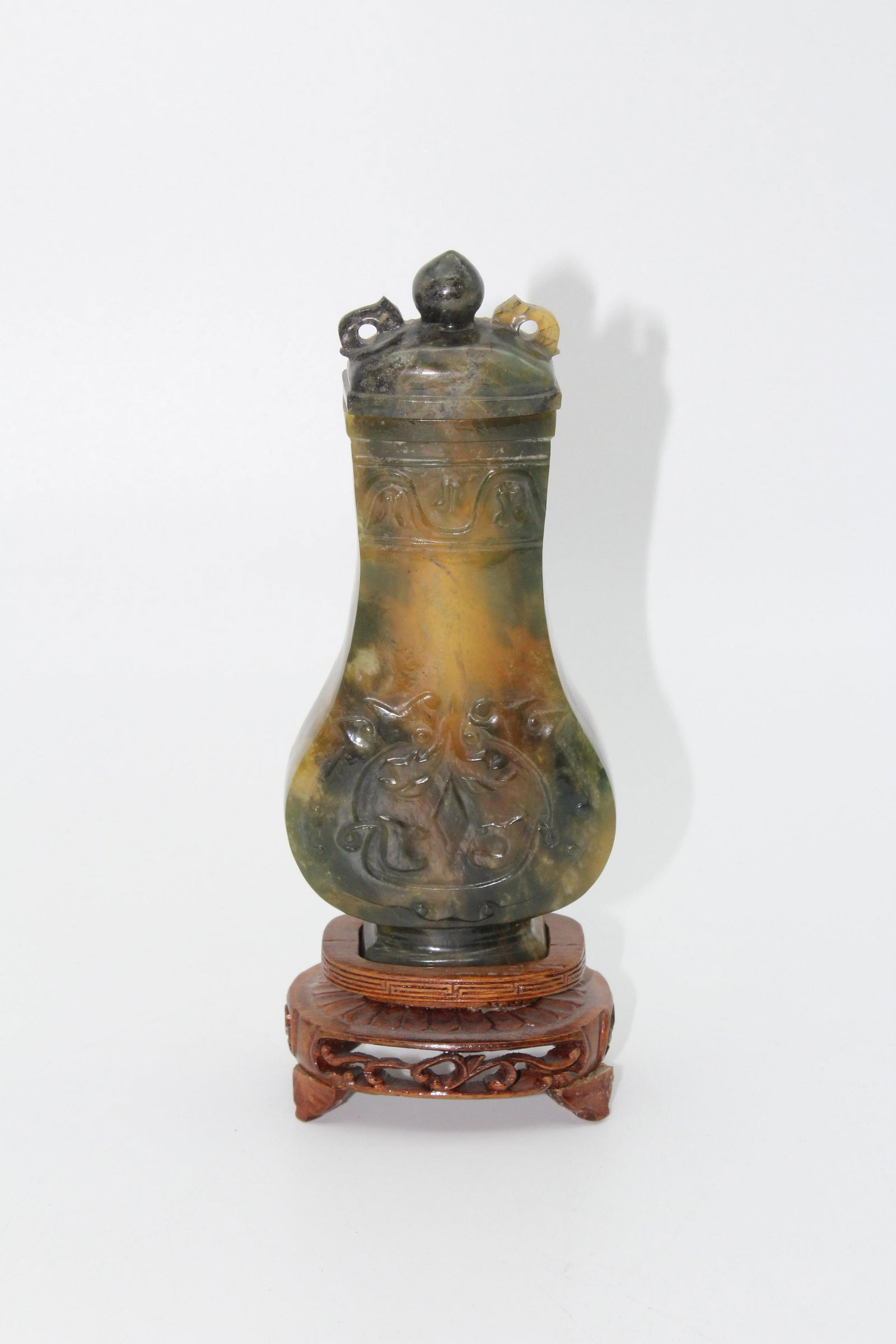 Carved Hardstone Covered Urn on Wooden Stand: Carved Hardstone Covered Urn on Wooden Stand, lid has double handles on it, carved design on body. Size: Urn: 6.75" H x 3" W x 1" D.