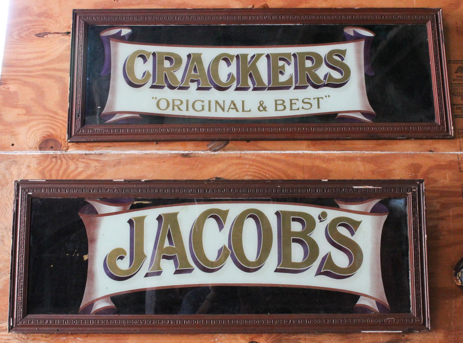 Lot of 2 Reversed Painted Glass Signs: A lot of two reversed painted glass English advertising signs. "Jacob's" & "Crackers Original & Best". Both in shadow font and framed. Embossed frames spell out Jacobs Biscuits. Size: 16"H x 44"W each