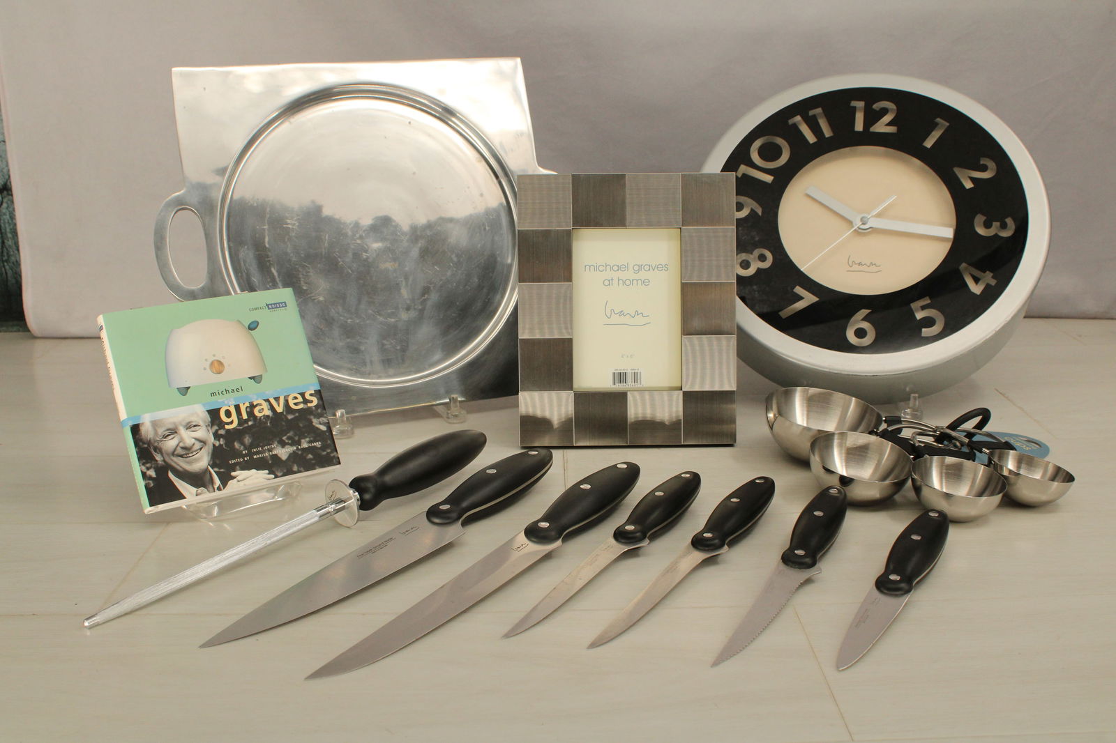 Generous Michael Graves design lot. Platter, knife set, picture frame, clock, book, measuring cups: Generous Michael Graves design lot. Platter, knife set, picture frame, clock, book, measuring cups and promotion posters. Sizes: Various, Tray 13.5"T x 16"W, book 6.25"T x 6.25"W, Knives 14"L-8.5"L, c
