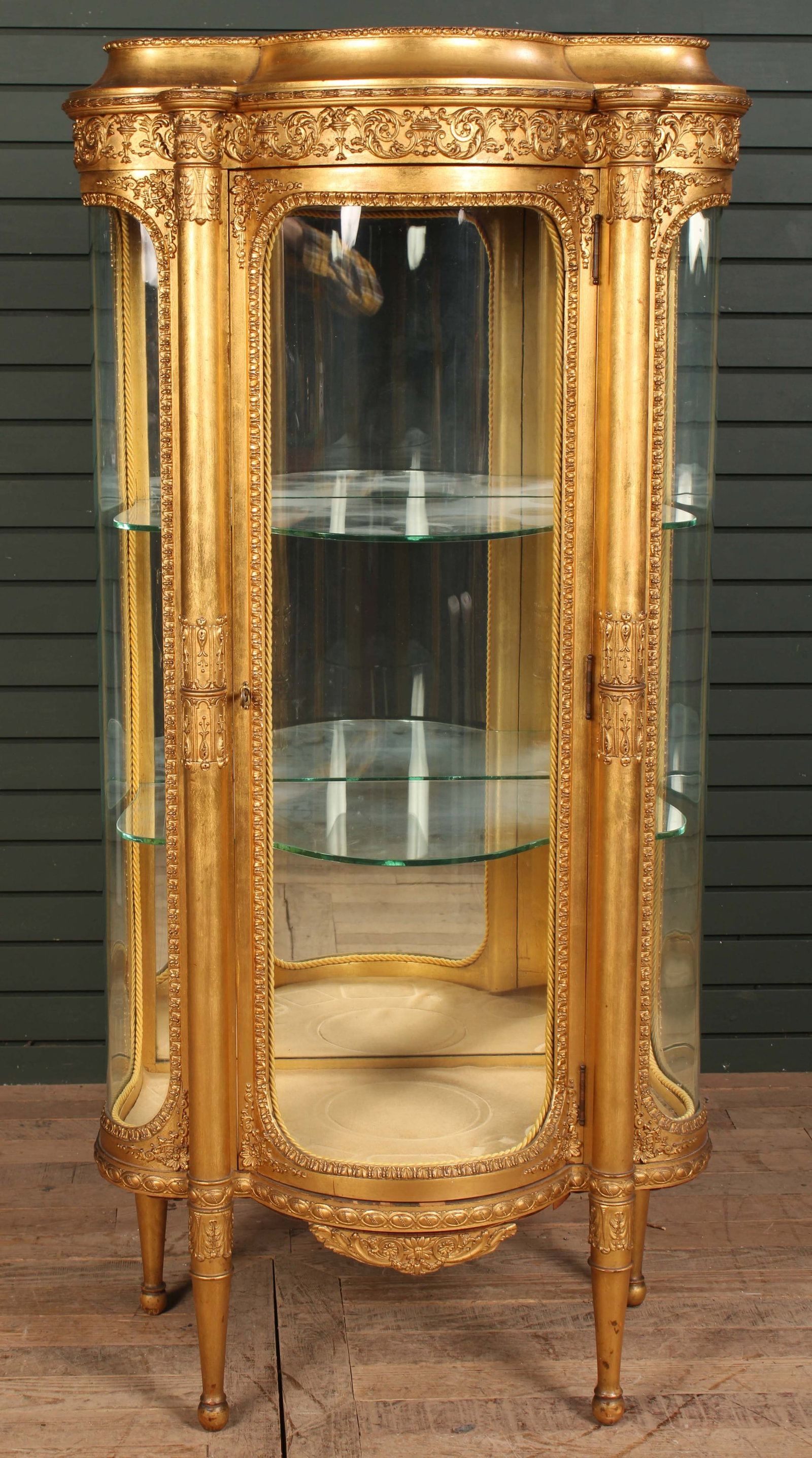 Gold Vitrine With Curved Glass Front: A Continental gilt wood carved Vitrine with stepped top over curved glass sides framed by decorated columns. Two glass shelves. Mirrored back. Size: 56.5"H x 28"W x 17"D