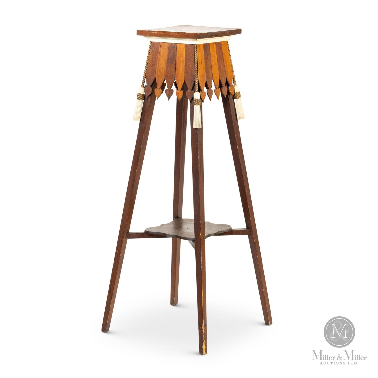 Fern Stand with Carved Corner Tassels: A tall fern stand with splayed legs and a square top with a carved white and gold tassel hanging from each corner and an apron consisting of light and dark wooden slats with spear tips pointing to the