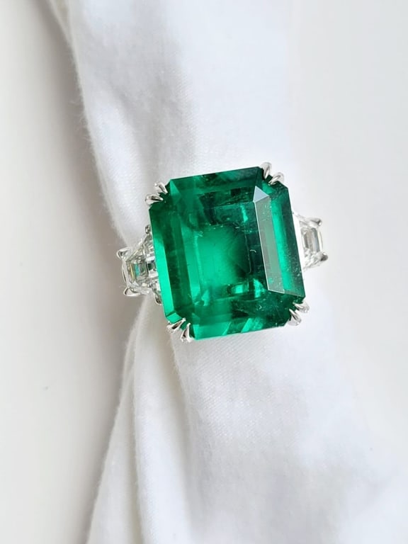 COLOMBIAN EMERALD RING WITH DIAMOND: A simply stunning and substantial (AGL and Gubelin Certified) Emerald is the star of this classic platinum ring. The 17.81 ct vibrant green Emerald is embraced on both sides by high quality trapezoid