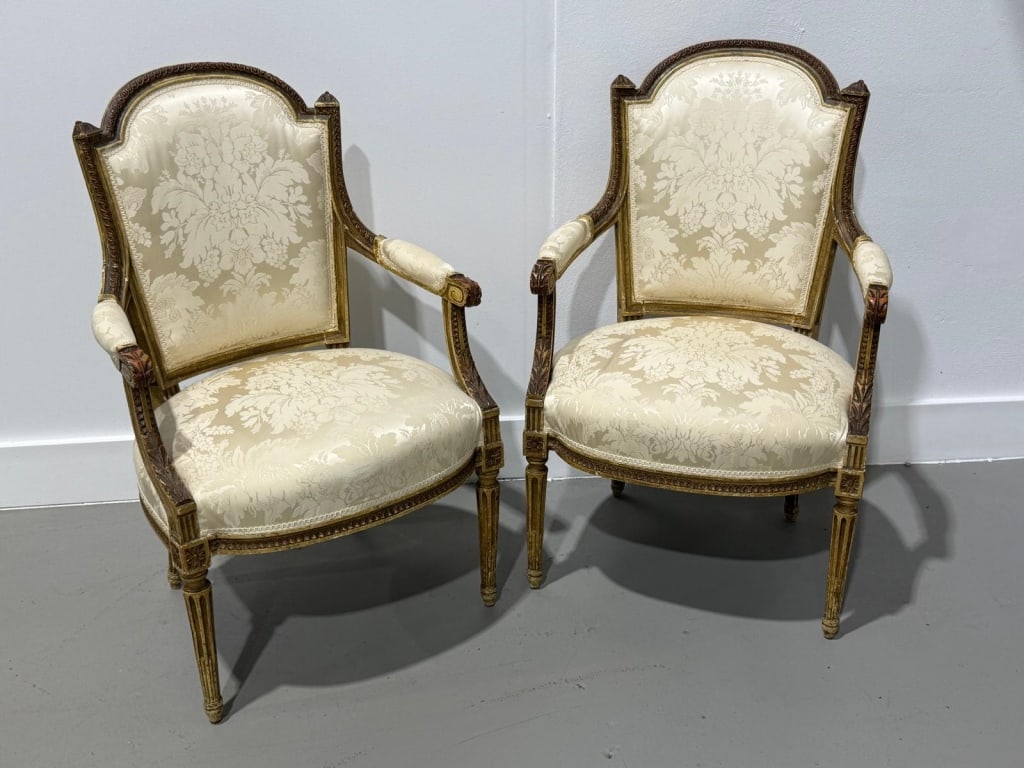 Pair of Louis XVI Style Upholstered Armchairs: Elegant pair of Louis XVI style fauteuil armchairs featuring carved and giltwood frames with shaped crest rails, fluted legs, and scrolling arms with padded rests. Upholstered in a cream floral damask