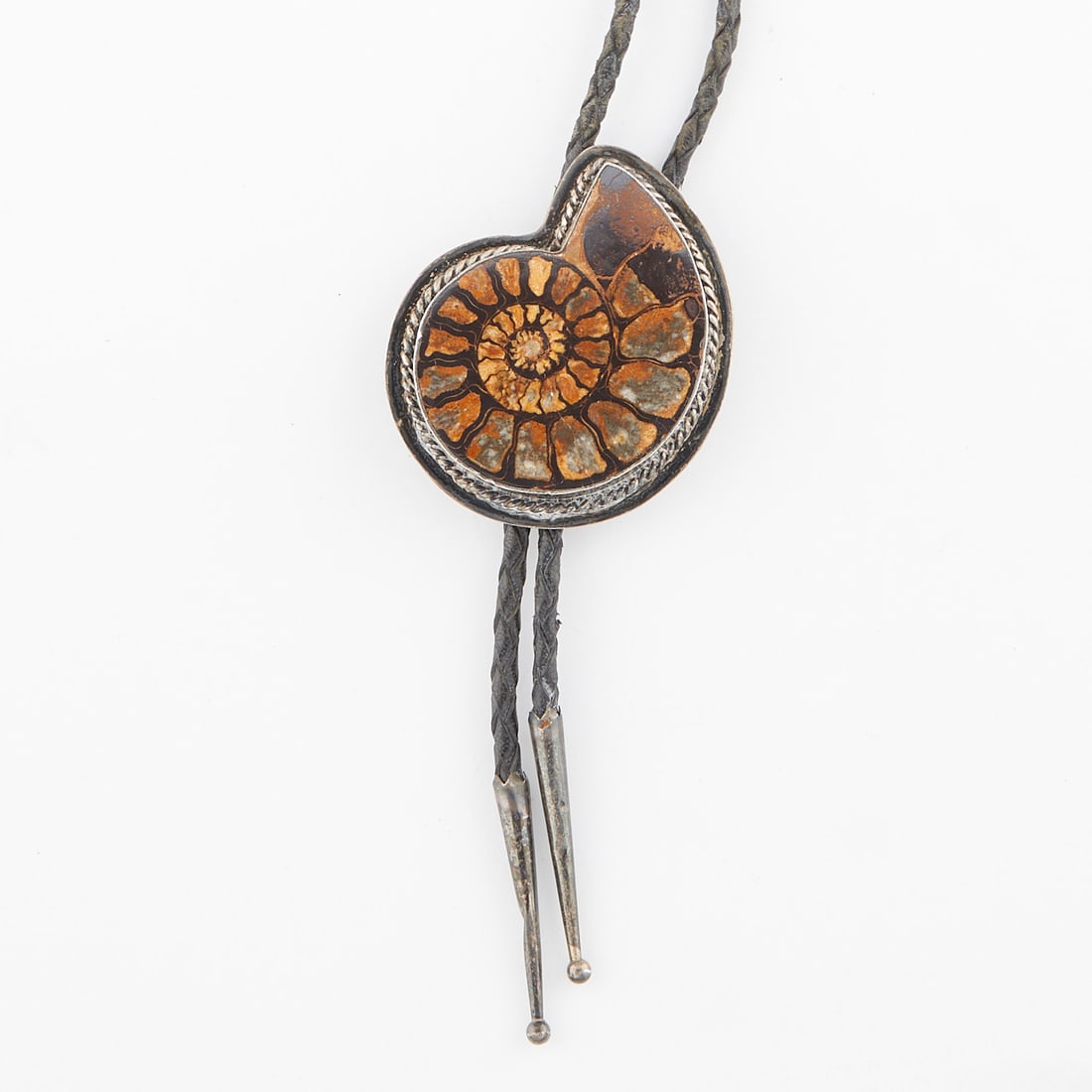 Silver & Ammonite Fossil Bolo Tie: Bolo tie with a clasp comprised of an ammonite fossil set silver with rope detail bezel, on black braided leather with silver aglets.Overall length: 22 in. Clasp width: 54.95 mm x height: 64.34 mm. Gr