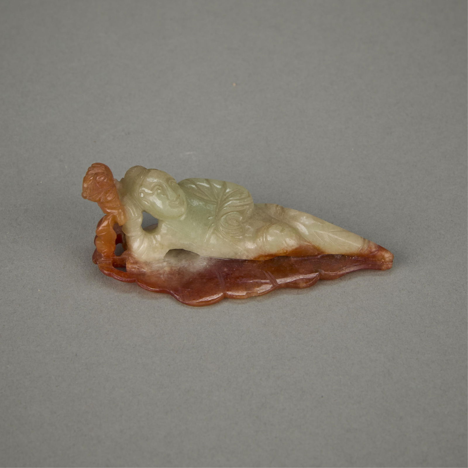 Chinese Carved Jade of a Reclining Figure on Leaf: Chinese carved jade depicting a figure reclining along the length of a leaf. The body is of a pale green coloration and the foliage is skillfully carved from a russet inclusion.Height: 1 in x width: 2