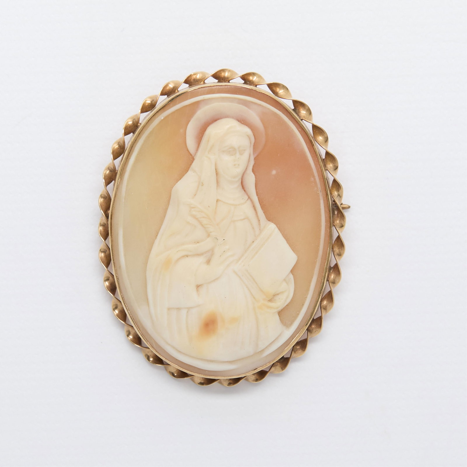 14k Cameo Brooch of St. Theresa of Avila: 14k yellow gold brooch set with a shell cameo depicting St. Theresa of Avila holding her attributes of a quill and book, in the habit of the Carmelite order. With a twisted frame and locking c clasp c