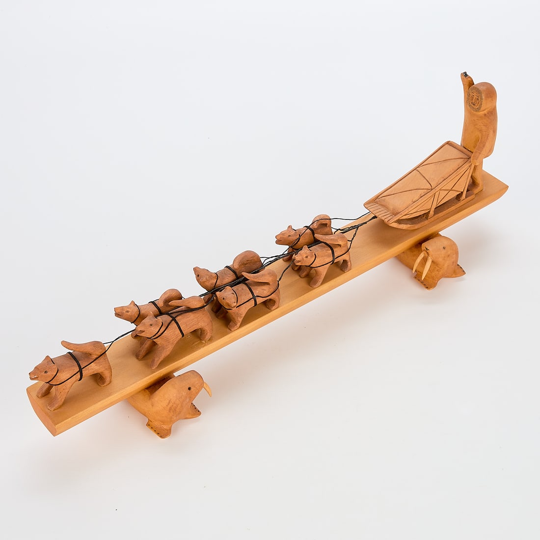 Paul Imana Wood Carved Sled Dog Scene: Paul Imana carved wooden sculpture depicting an inuk and sled dog team comprised of a sled and seven dogs. Mounted on a rectangular base supported by two walruses. Signed Paul Imana in ink along the u